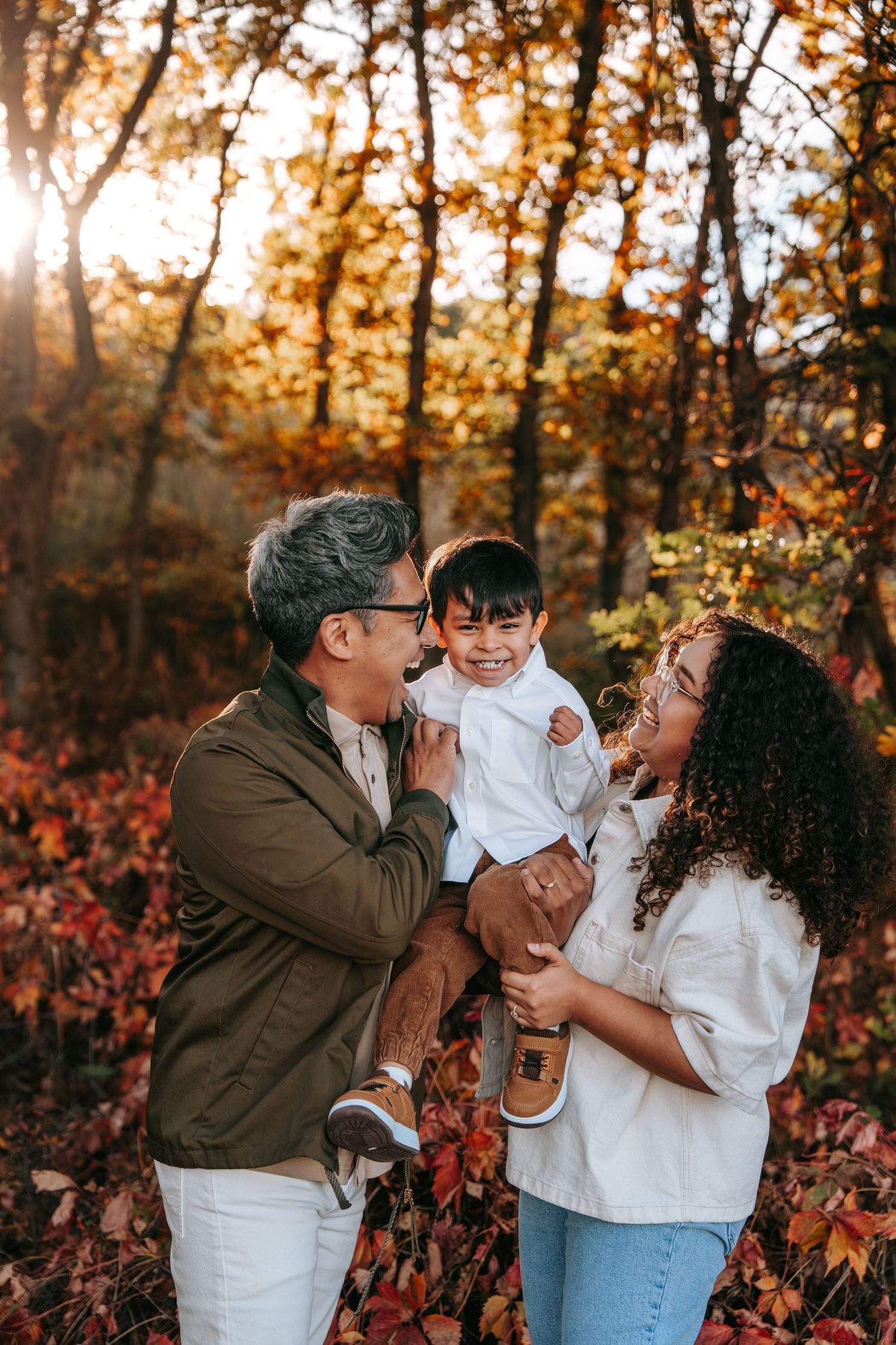 Joyful interracial family of three laughing together in autumn forest with colorful fall foliage.