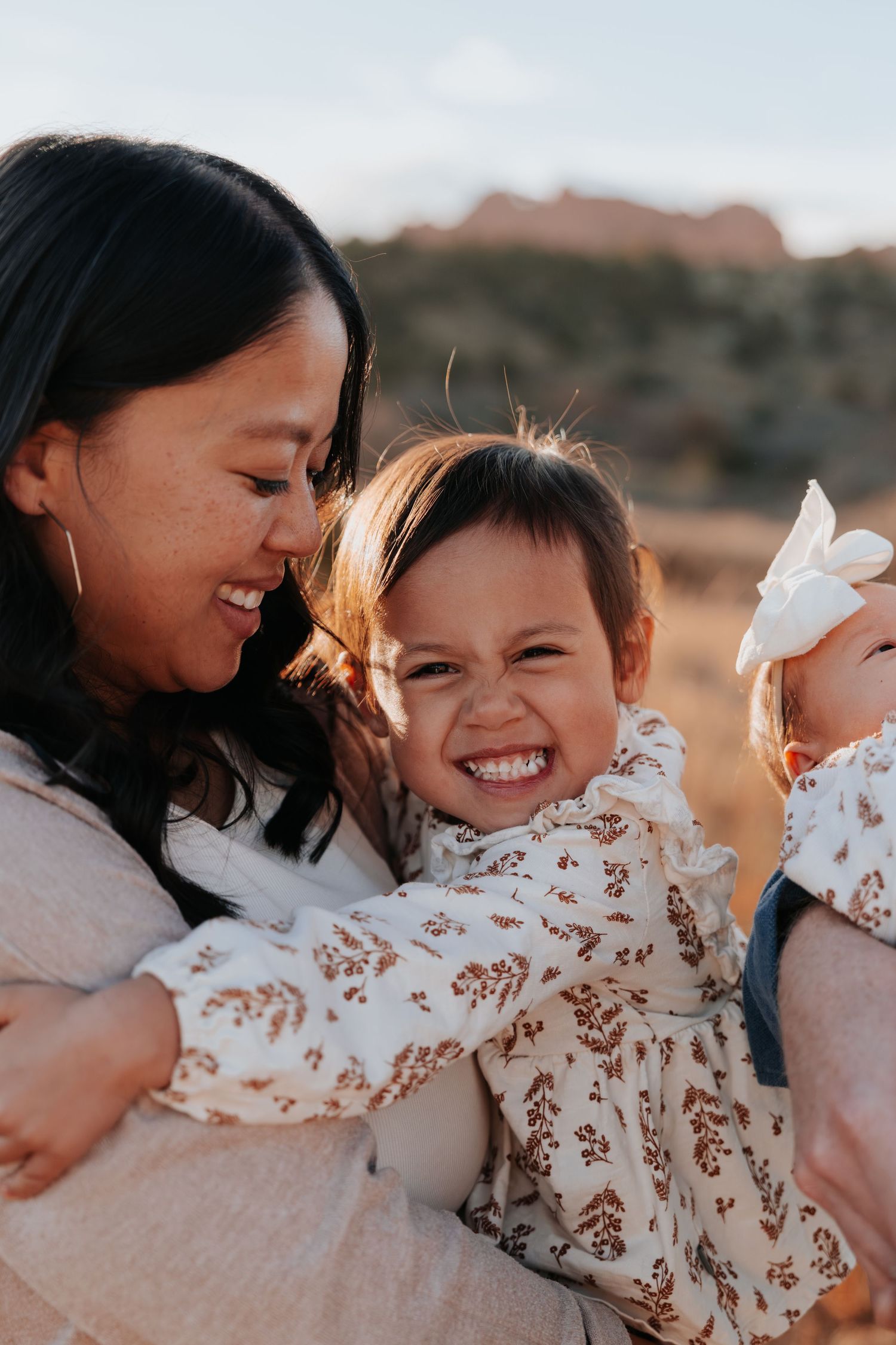 Mother laughing joyfully while holding two young daughters outdoors in warm sunset light.