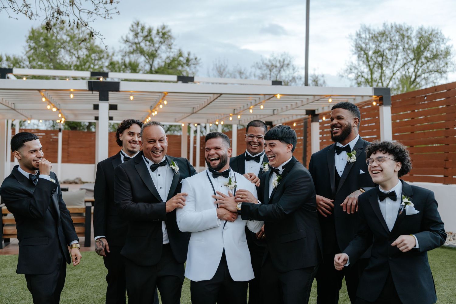 Groom in white tuxedo jacket laughs with groomsmen in black suits outdoors at elegant wedding venue.