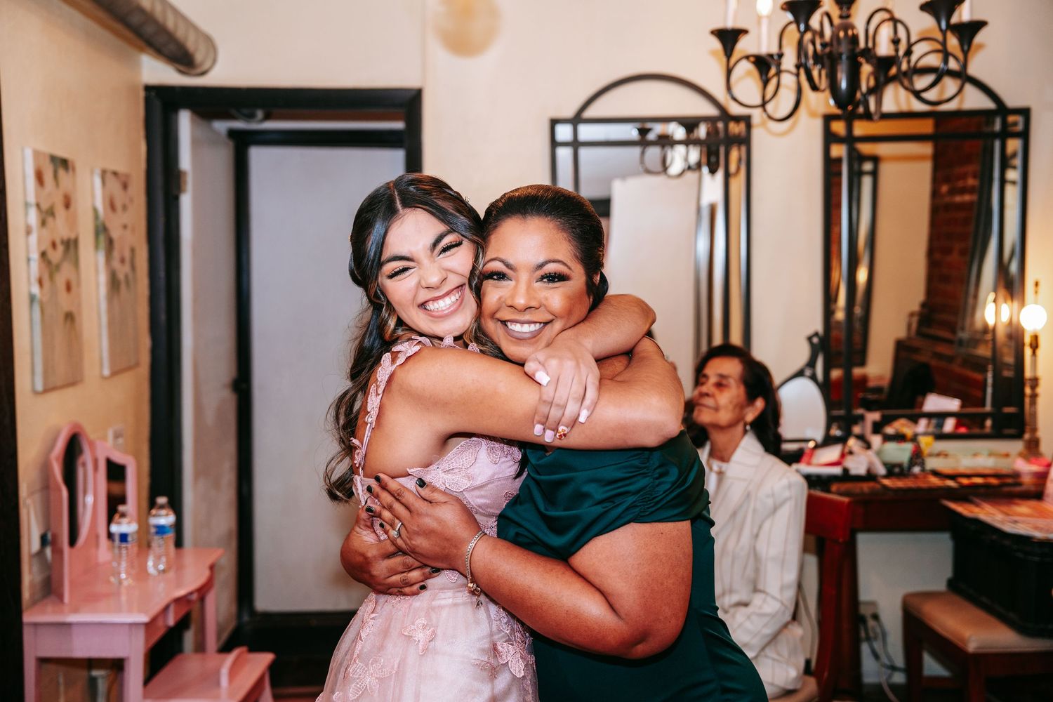 Quinceañera girl in pink gown sharing a warm hug with woman in green dress during celebration.