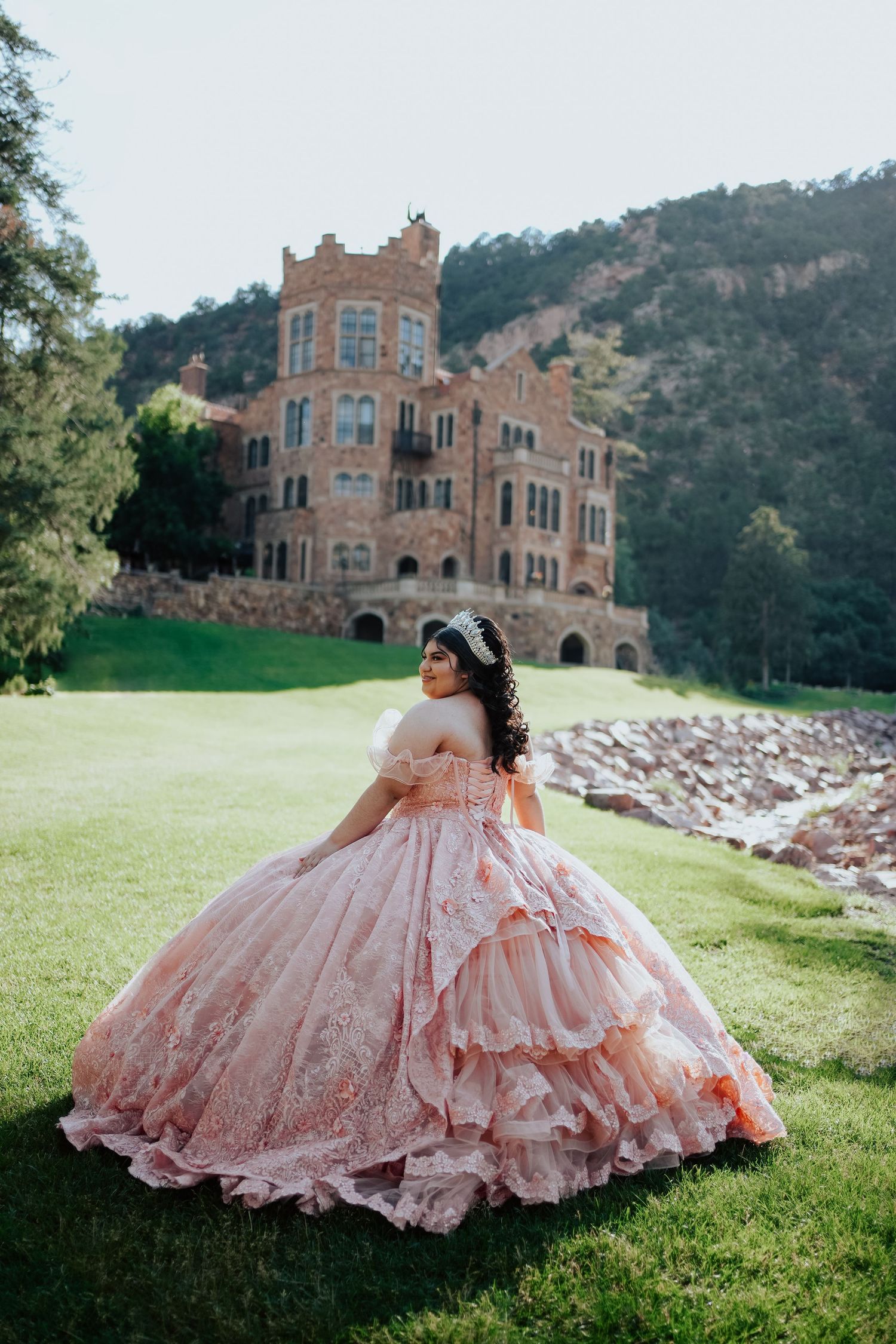 A young woman in a stunning pink layered quinceañera ball gown poses before a historic castle.