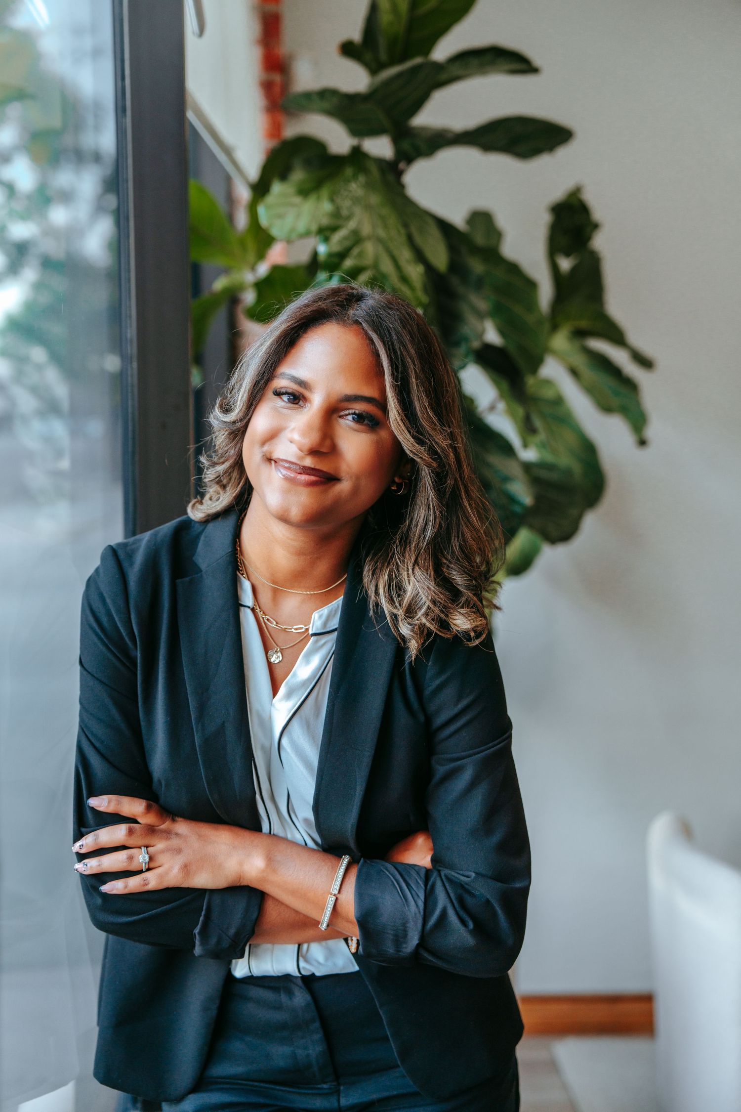 Poised businesswoman in black blazer standing with arms crossed beside a large green plant near a window.