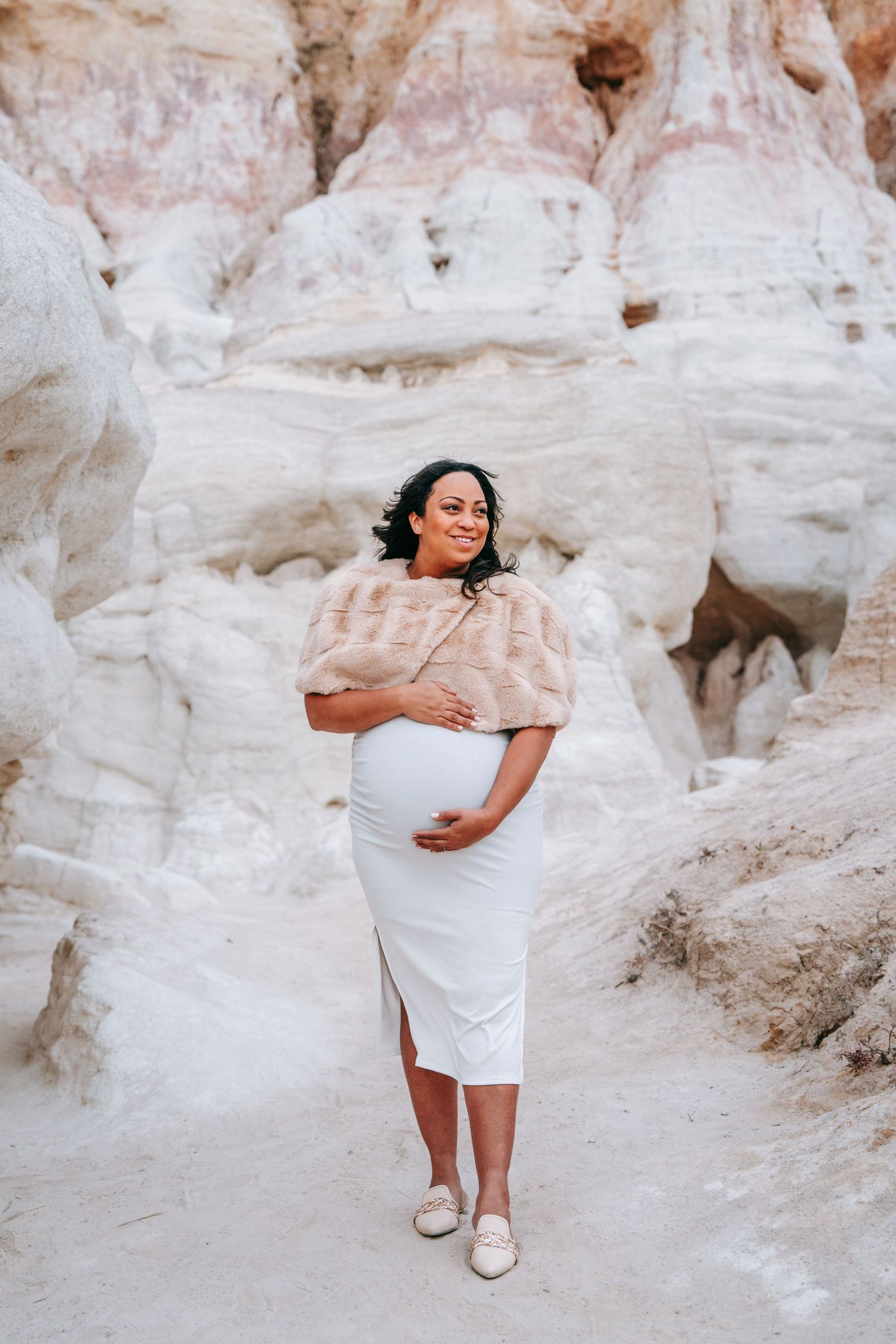Smiling pregnant woman in white dress and fur shawl posing in front of white rock formations.