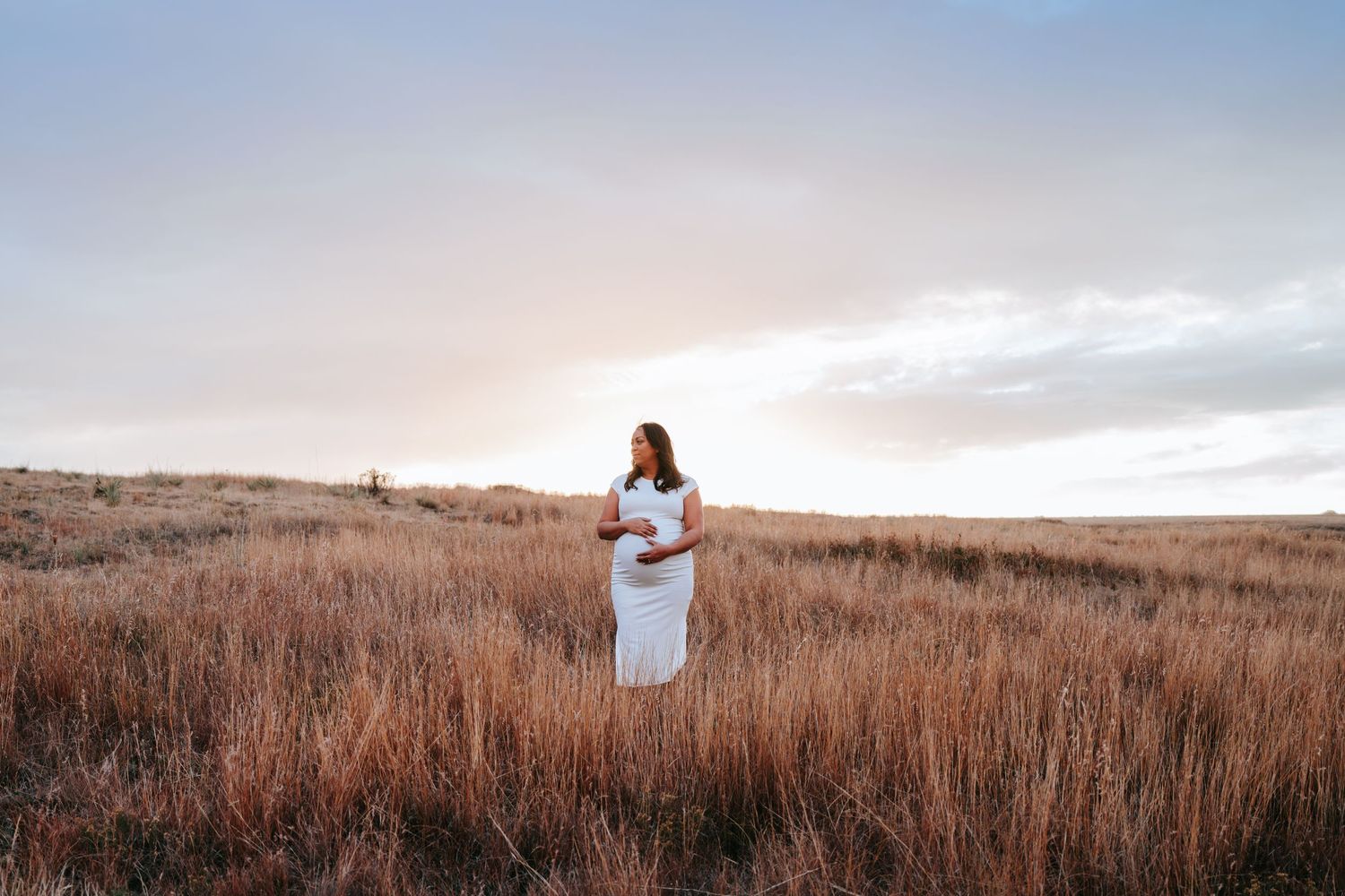 Pregnant woman in a white dress standing in a golden field at sunset.