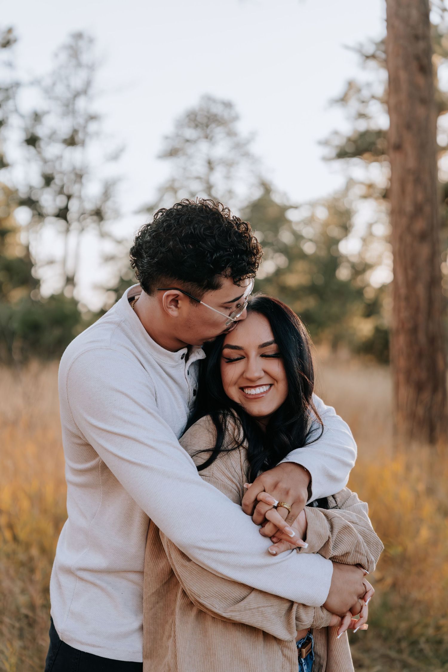Man kisses woman on the cheek while embracing her from behind in a sunlit autumn forest clearing.