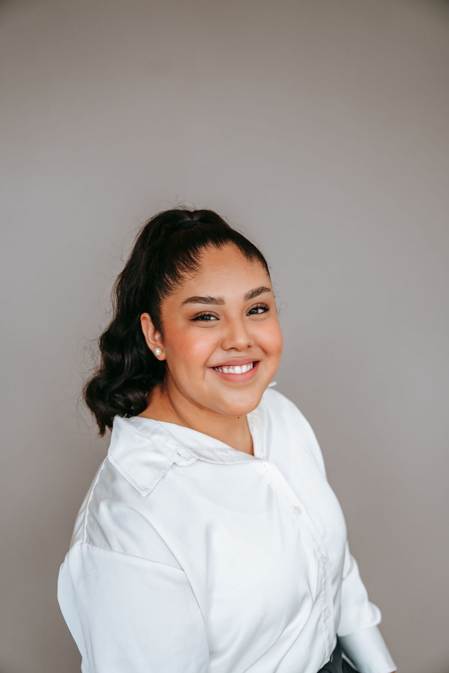 A young woman with dark hair smiles confidently in a white button-up shirt against a gray background.