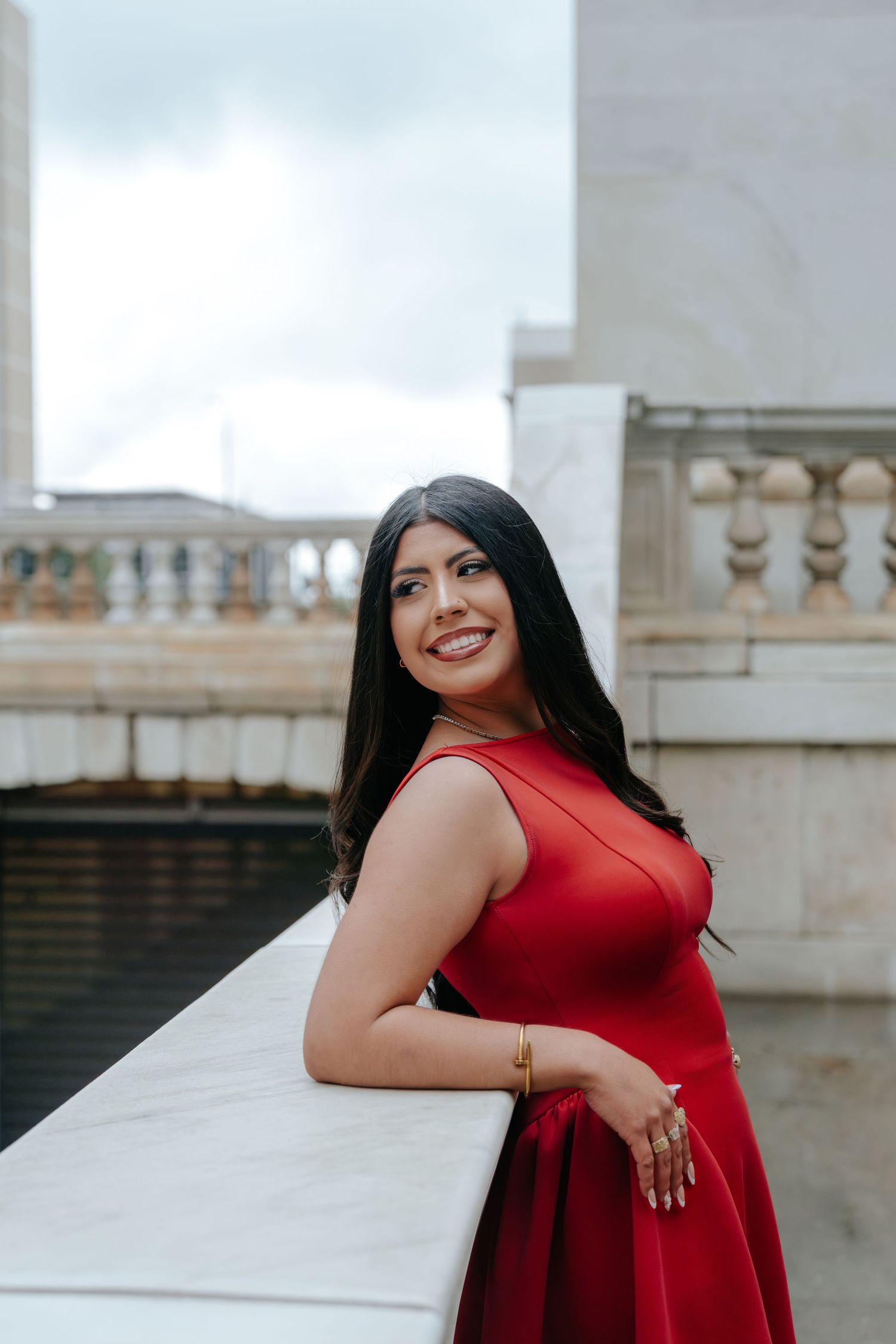 Smiling woman in red dress leaning on marble railing with classical architecture background.