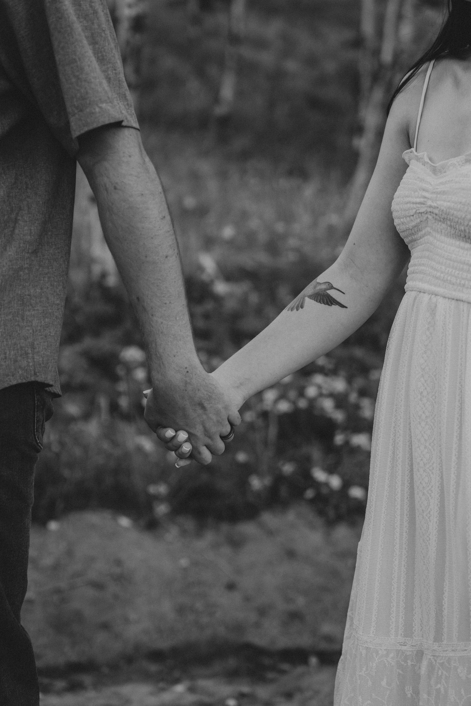 Black and white photo of a couple holding hands, woman with bird tattoo on forearm wearing white dress.