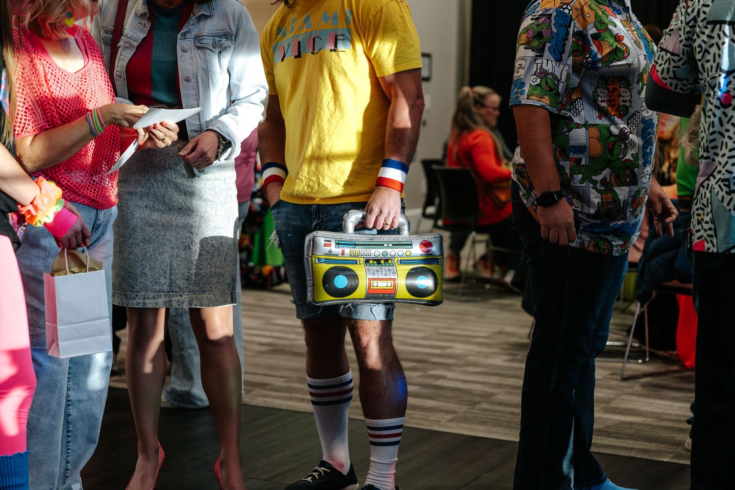 Person in yellow shirt and shorts holding inflatable boombox at a retro-themed 80s costume party event.