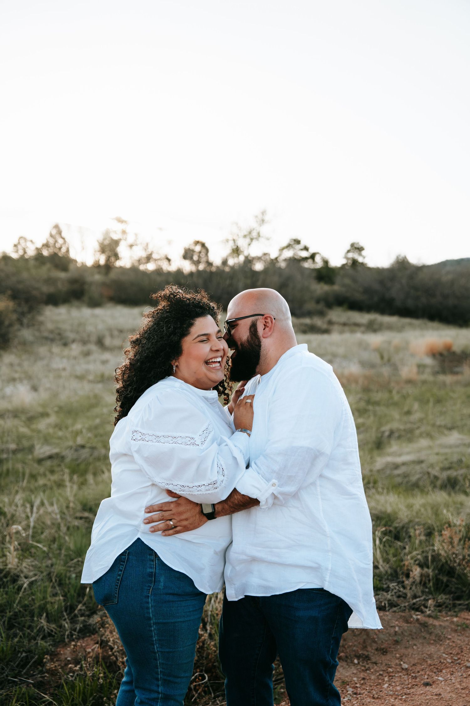 Happy couple in white shirts laughing and embracing closely on a dirt path in a grassy meadow.