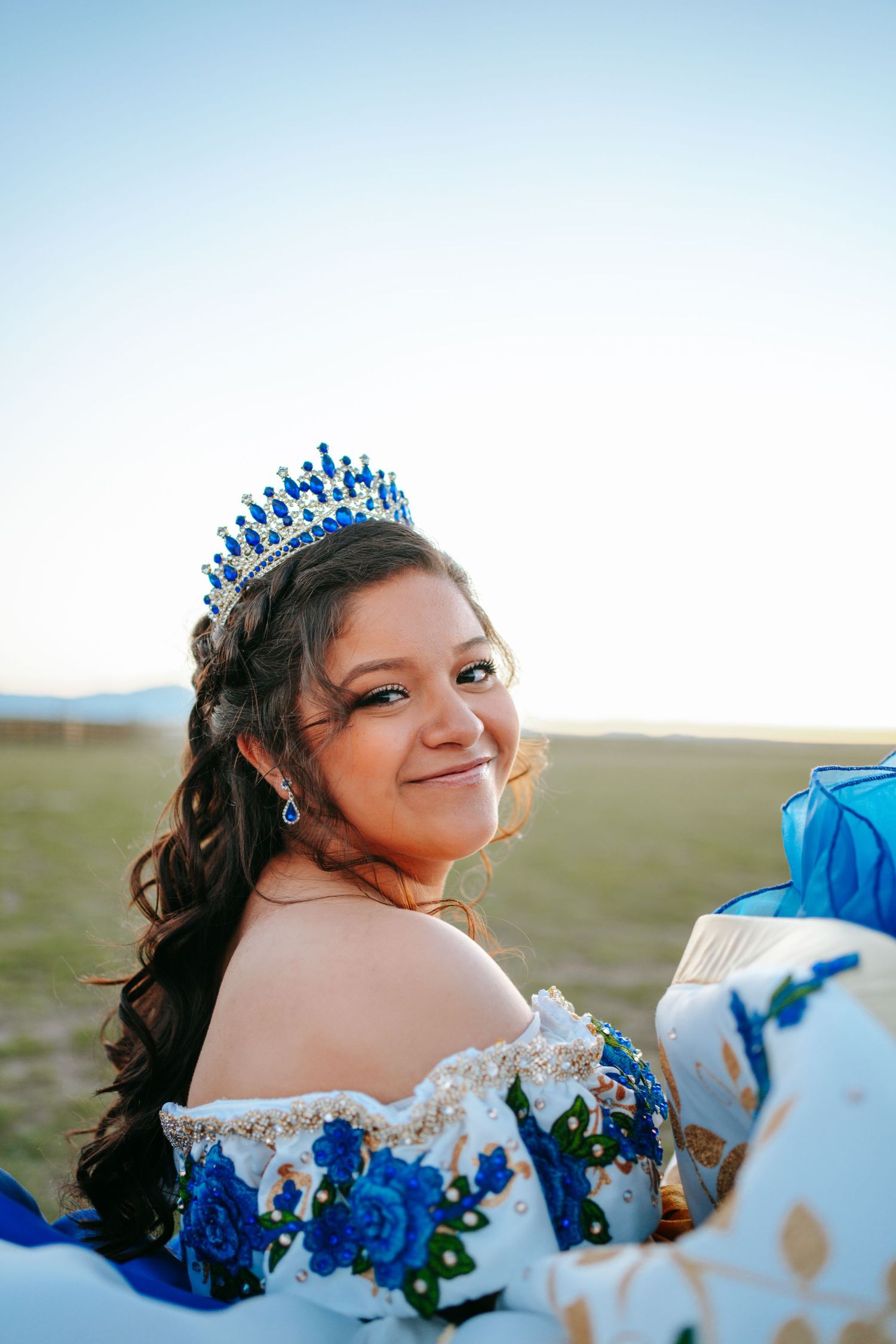 Smiling quinceañera wearing blue crystal tiara and floral embroidered white and blue gown outdoors.