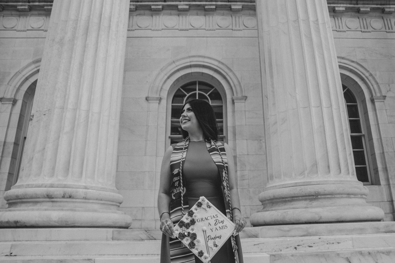 Black and white photo of smiling woman holding decorated graduation cap between stone columns.