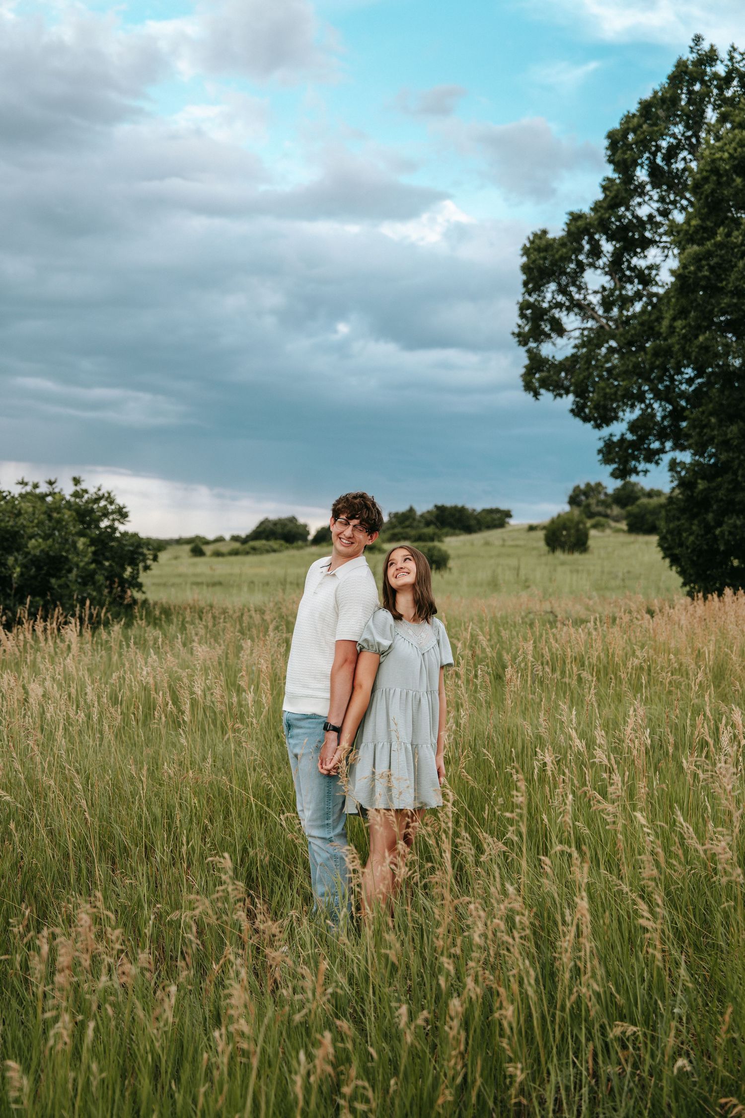 Happy couple standing in tall golden grass field under dramatic cloudy sky near a tree.