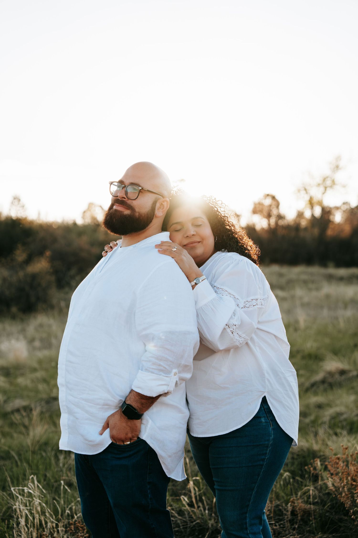 Couple in white shirts embracing at sunset in a grassy field, woman resting head on mans back.