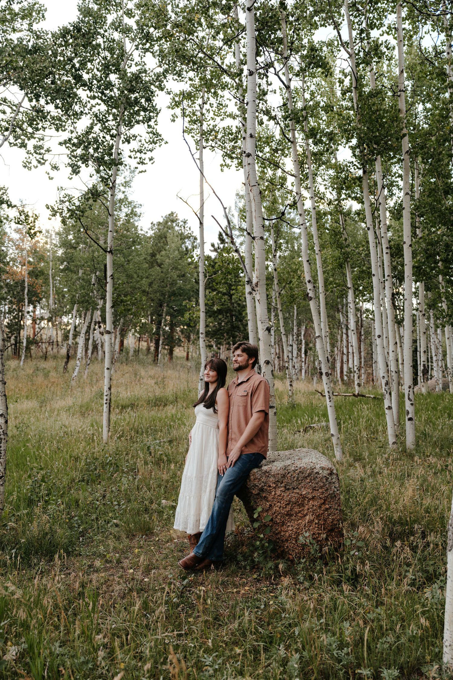 Couple standing by a rock in an aspen forest, woman in white dress, man in brown shirt and jeans.