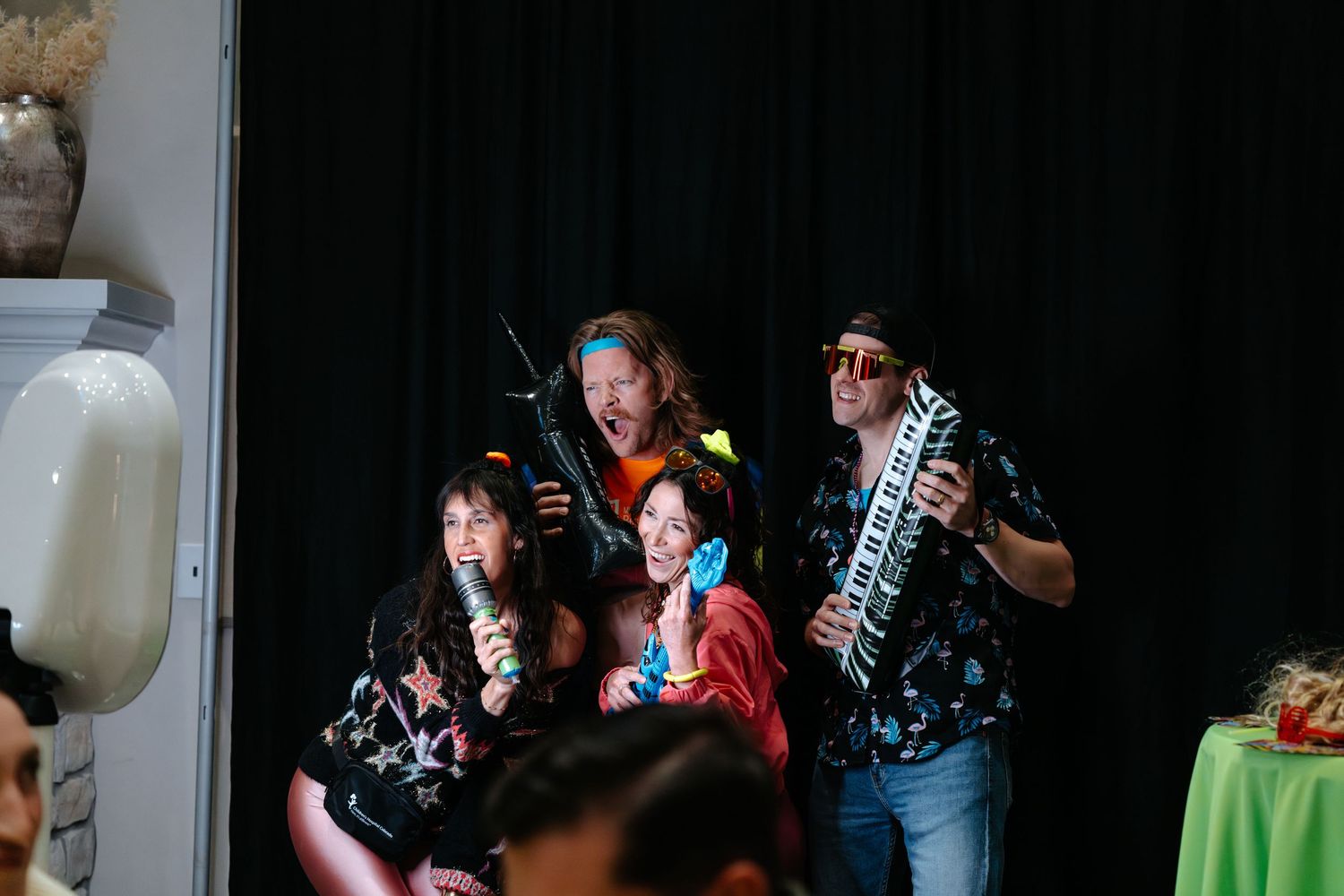 Four performers in Hawaiian shirts pose with instruments and microphone on stage before a dark curtain backdrop.