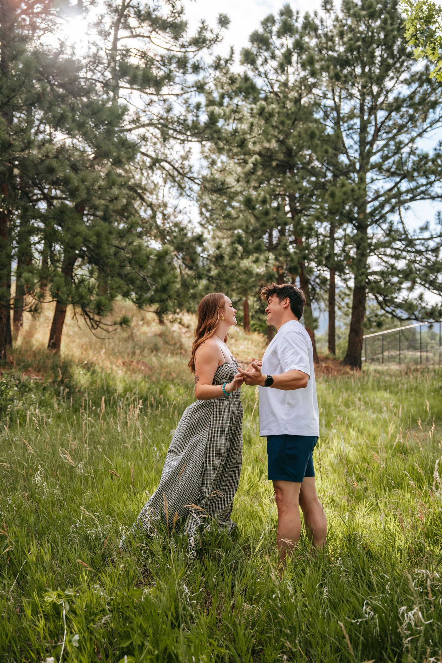 Couple holding hands in a sunlit mountain meadow surrounded by tall pine trees and lush green grass.