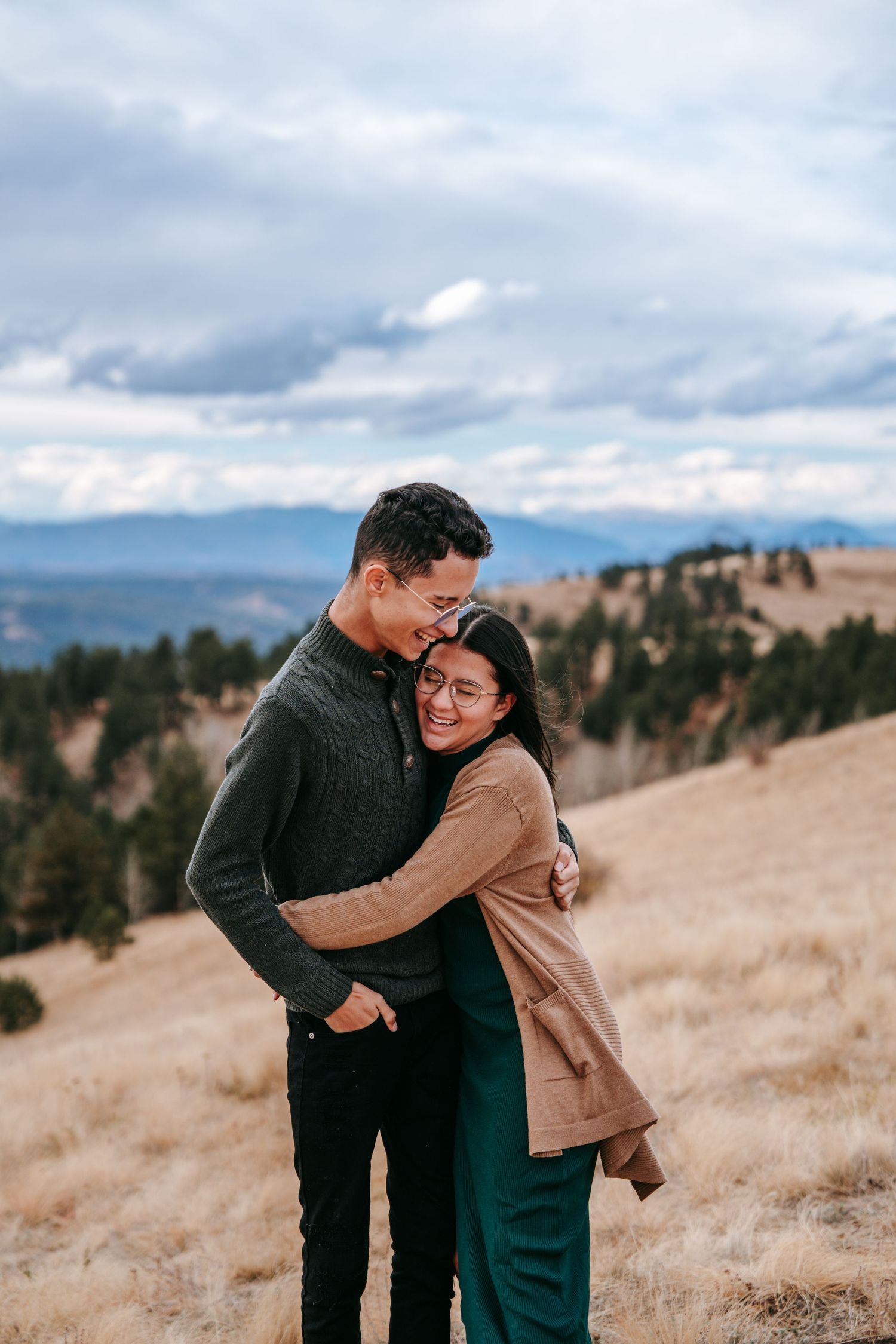 Young couple embracing warmly in a golden mountain meadow with scenic mountain views behind them.