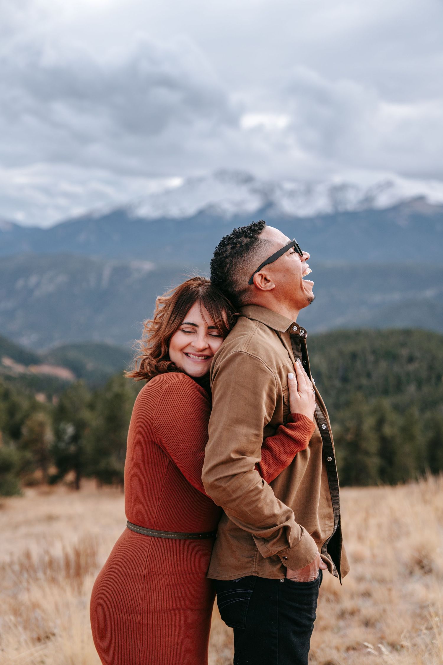 Woman in rust dress hugging man from behind with snowy mountain backdrop.