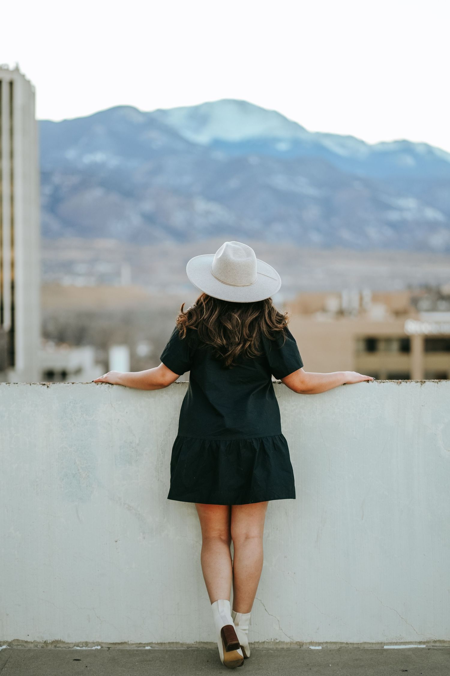 Woman in black dress and white hat gazing at mountain views from a rooftop.