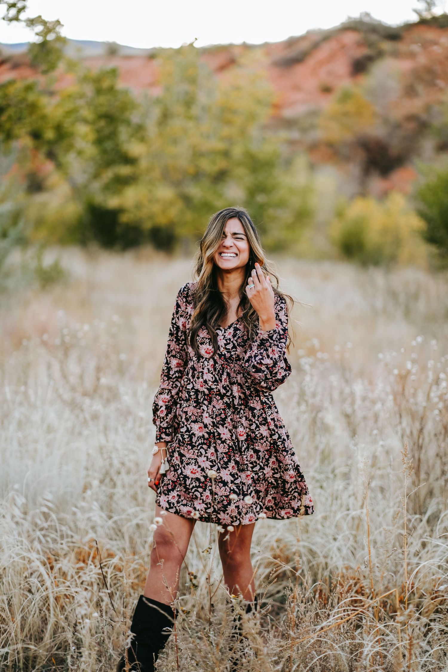 Young woman in floral dress and boots laughing in golden field with red rock landscape.