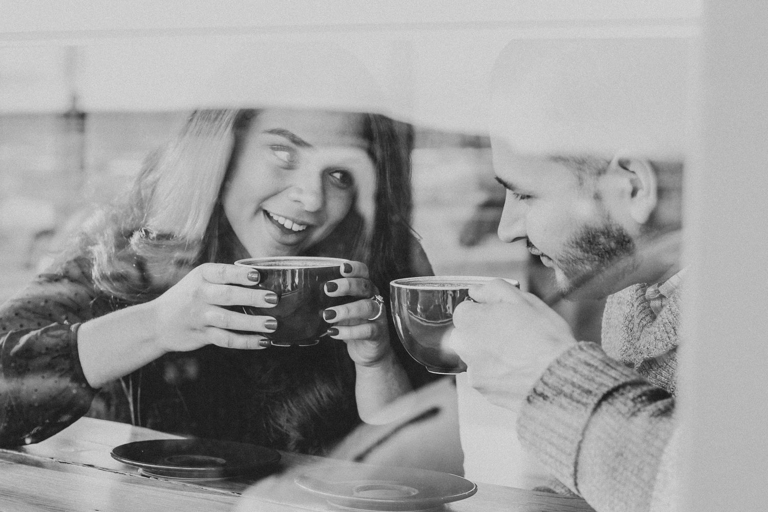 Black and white photo of couple laughing and sharing coffee mugs by a window.