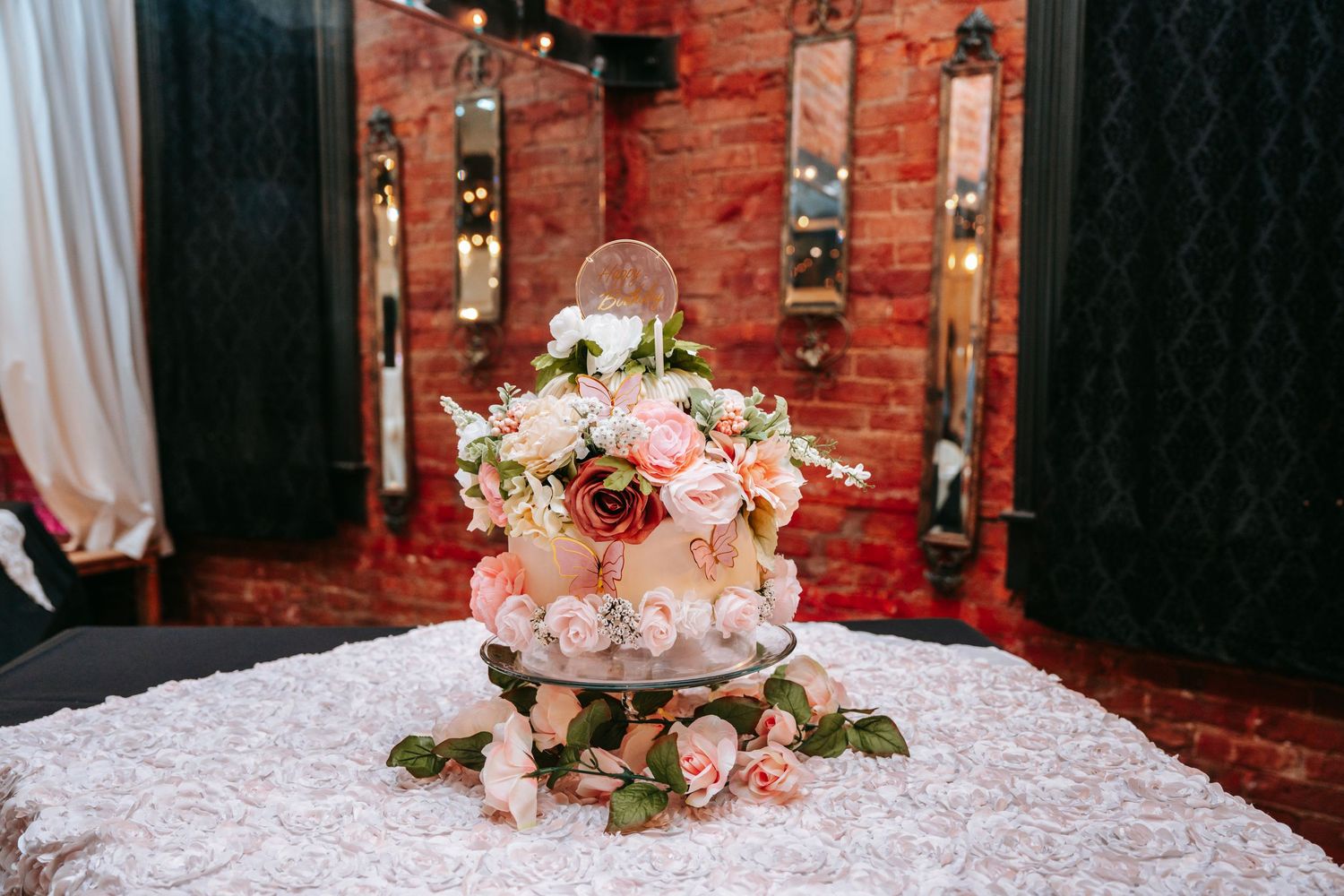 Elegant two-tier wedding cake adorned with pink roses and a dreamcatcher topper on a floral tablecloth.