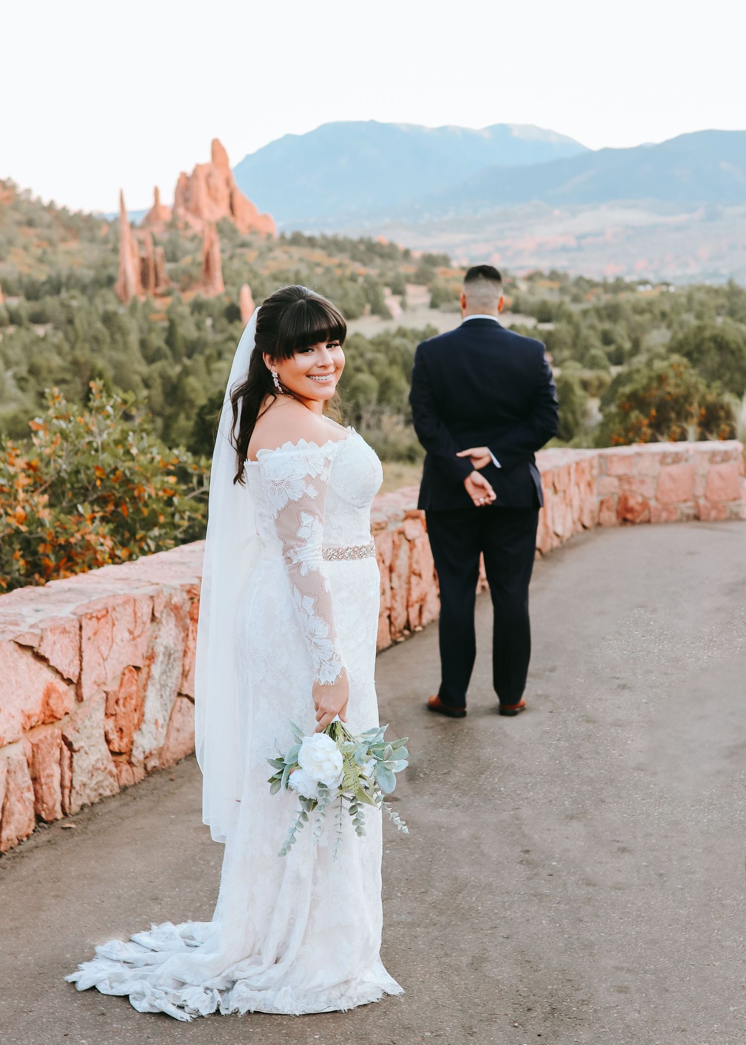 Bride in lace gown holding white bouquet with mountain and red rock scenic backdrop.