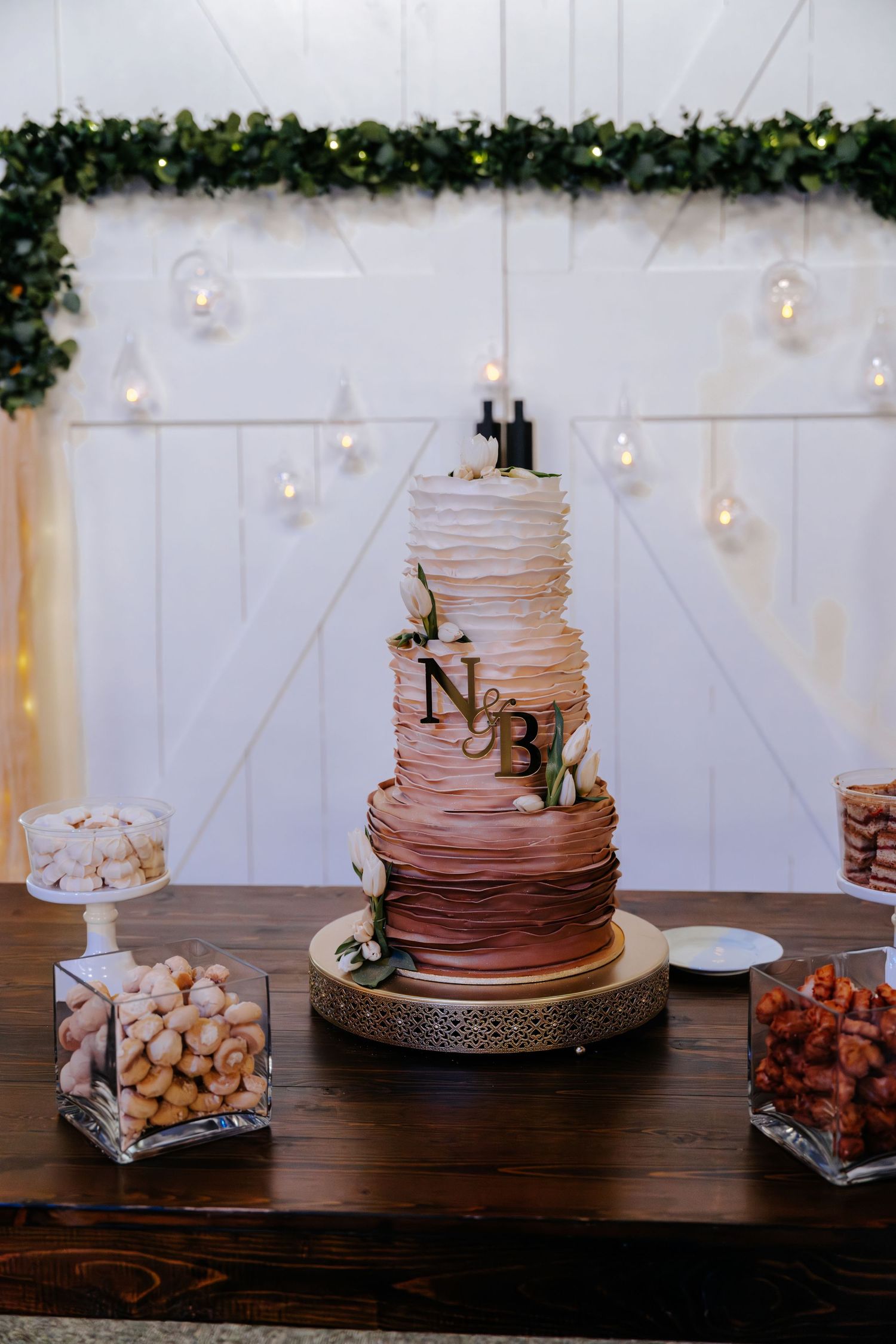 Elegant three-tier ombre pink ruffled wedding cake with gold monogram initials on a rustic wooden table.