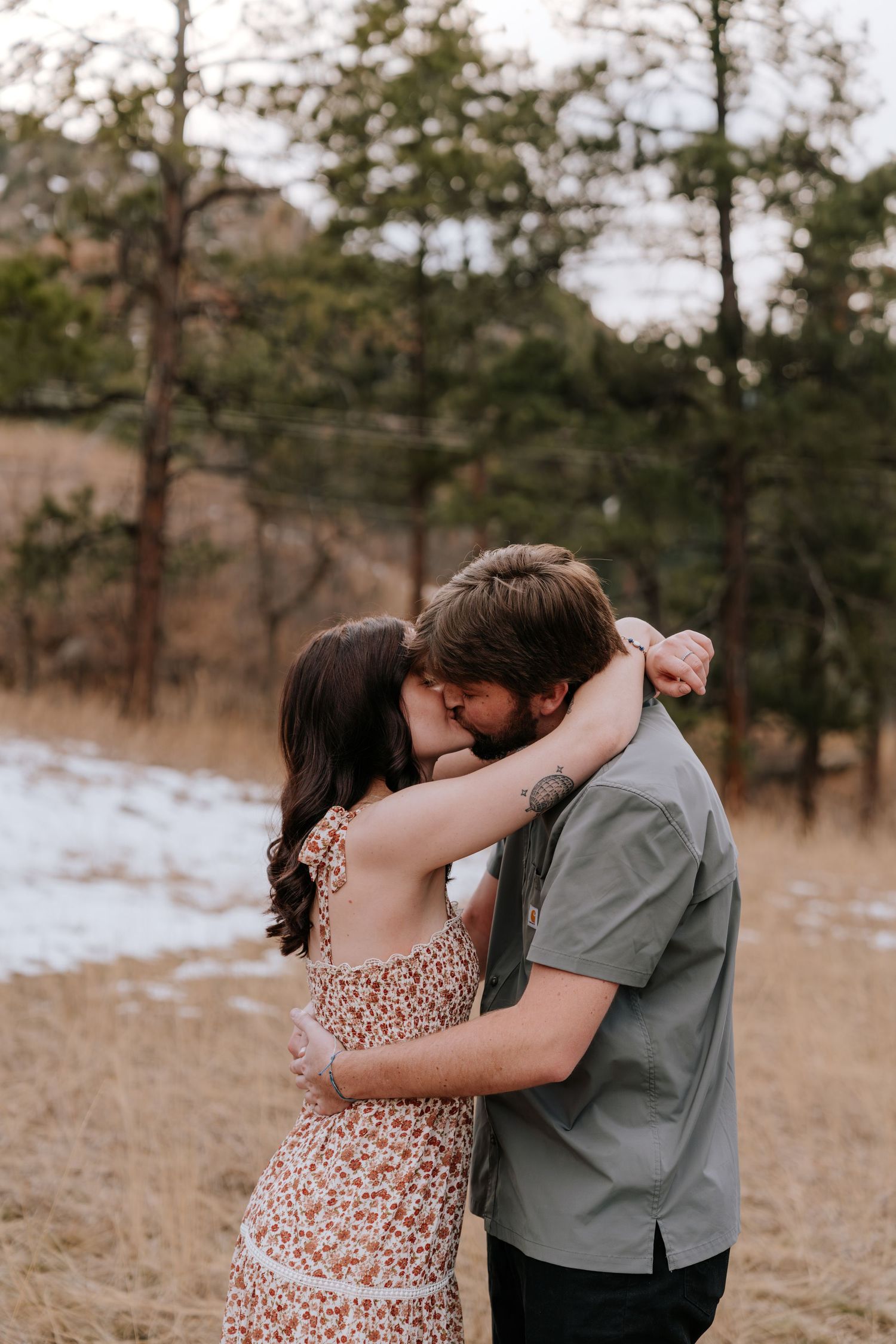 Couple sharing a romantic kiss in a snowy Colorado forest, woman in floral dress with arms around mans neck.