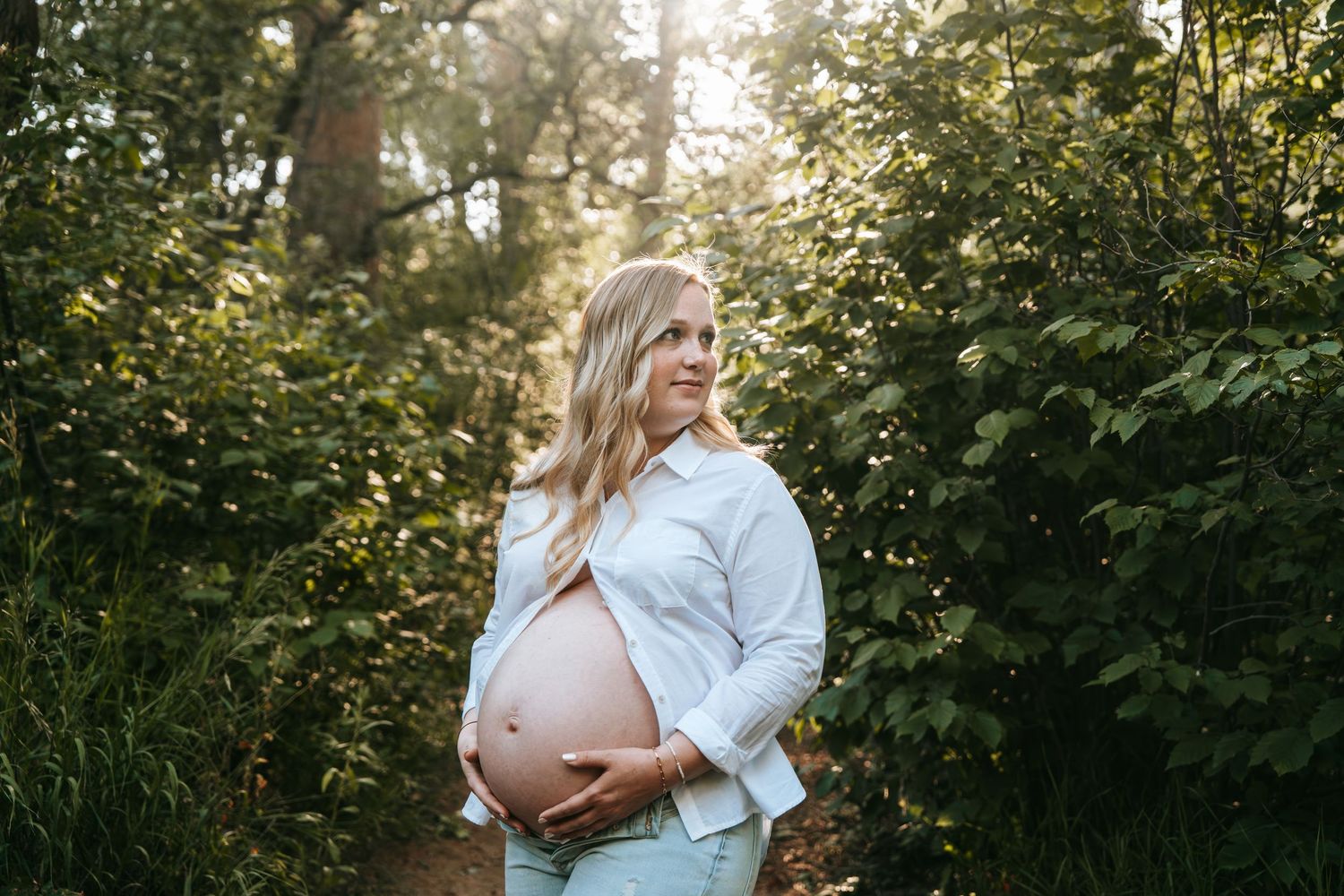 Pregnant blonde woman holding baby bump on a sunlit forest path surrounded by green foliage.