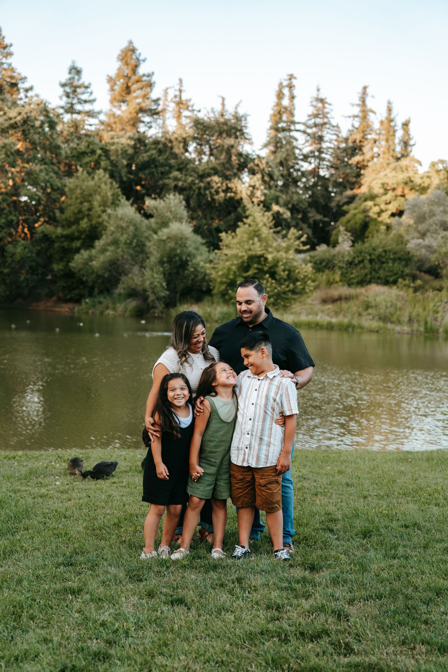A happy family of five stands together by a peaceful pond surrounded by tall evergreen trees.