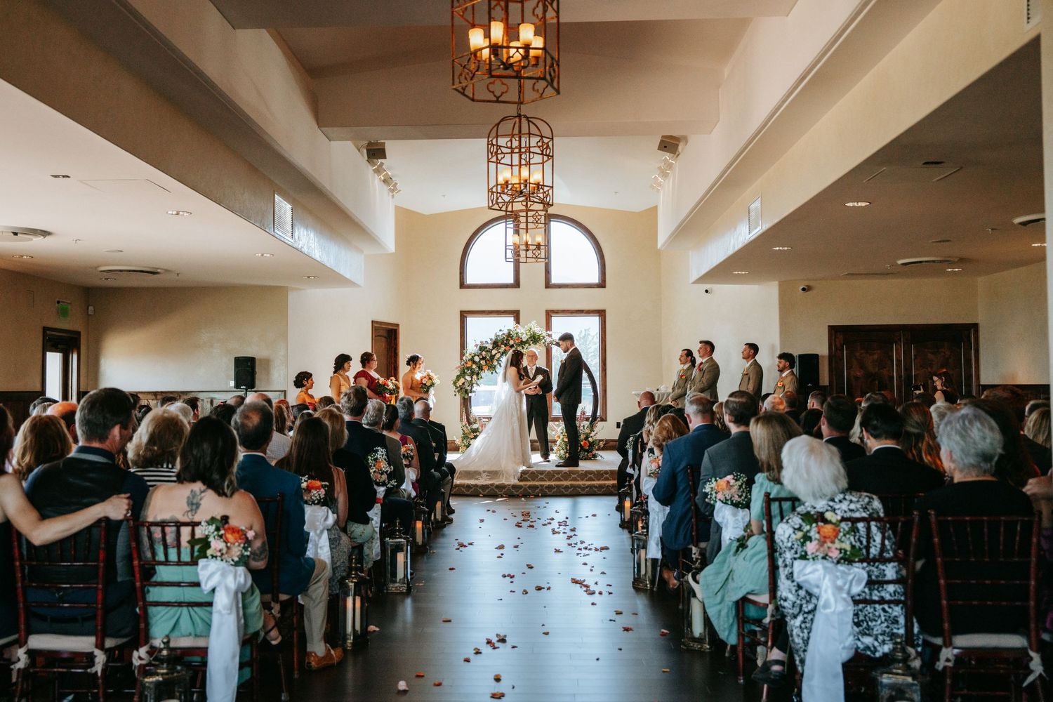 Wide-angle view of indoor wedding ceremony with couples at floral arch altar and guests seated in aisles.