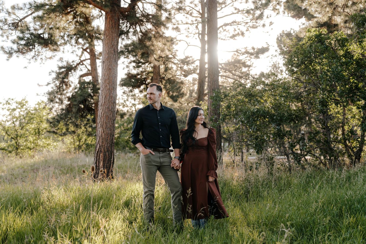 Couple holding hands in a sunlit forest meadow, woman in burgundy dress, man in black shirt.