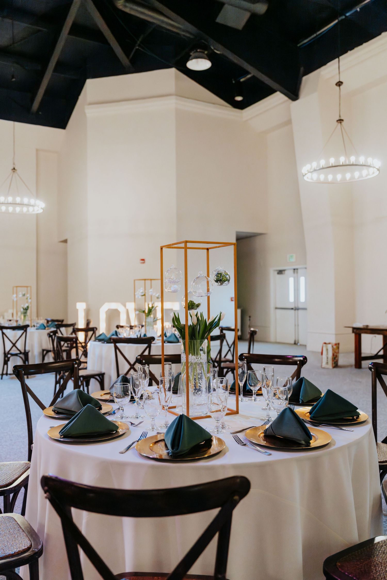 Elegant wedding reception hall with round tables, white linens, green napkins, gold chargers, and a gold geometric floral centerpiece.