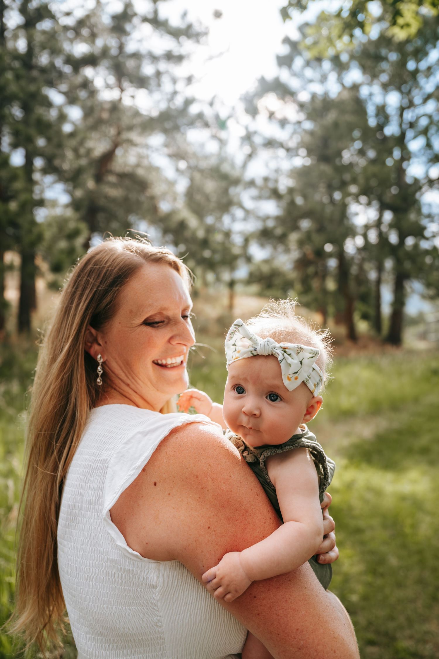 A smiling woman in white holds a baby girl with a bow headband outdoors in a sunlit forest setting.