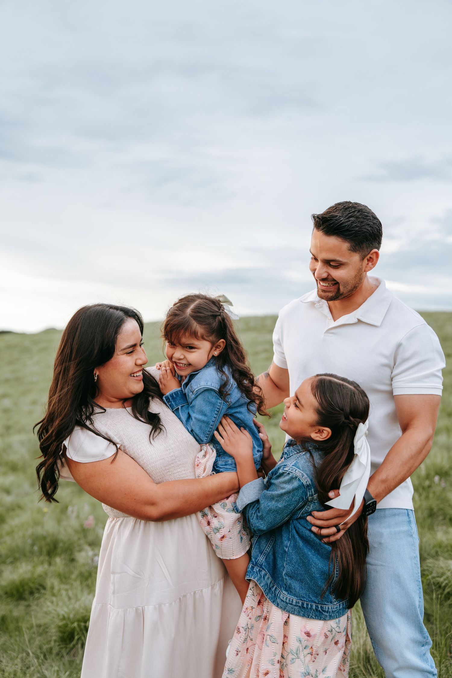 Happy family of four laughs together in a lush green meadow under a cloudy sky.