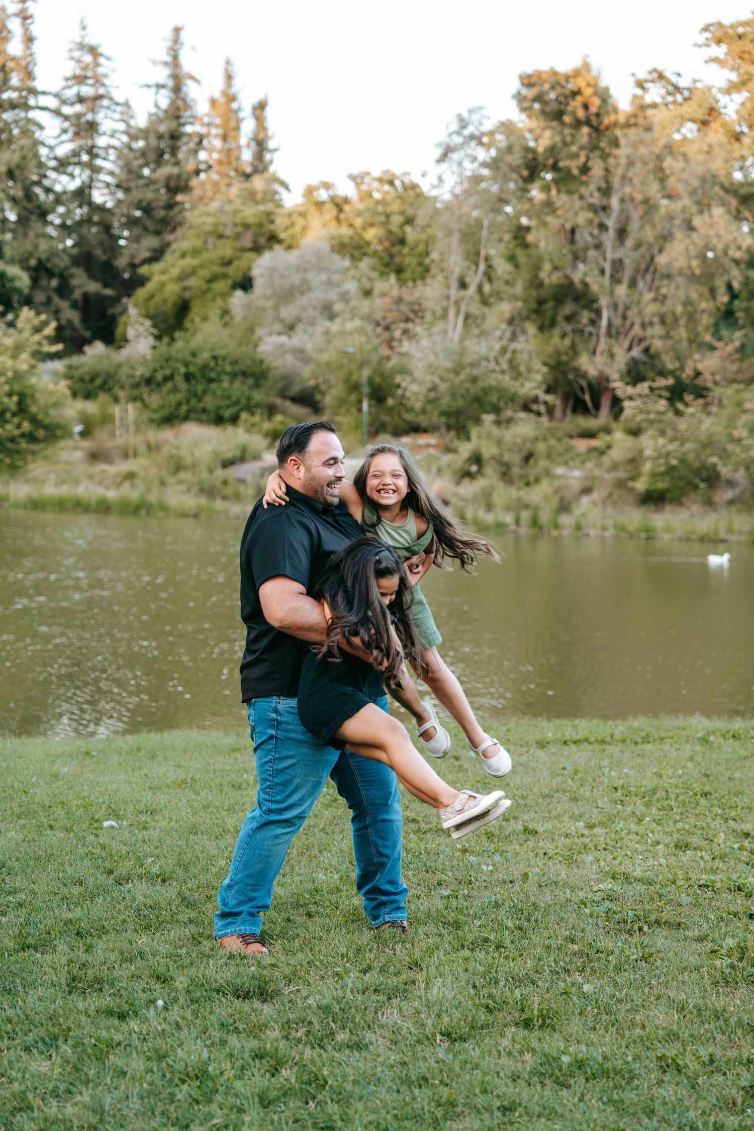 Father joyfully spinning his laughing daughter in the air beside a scenic park pond at sunset.