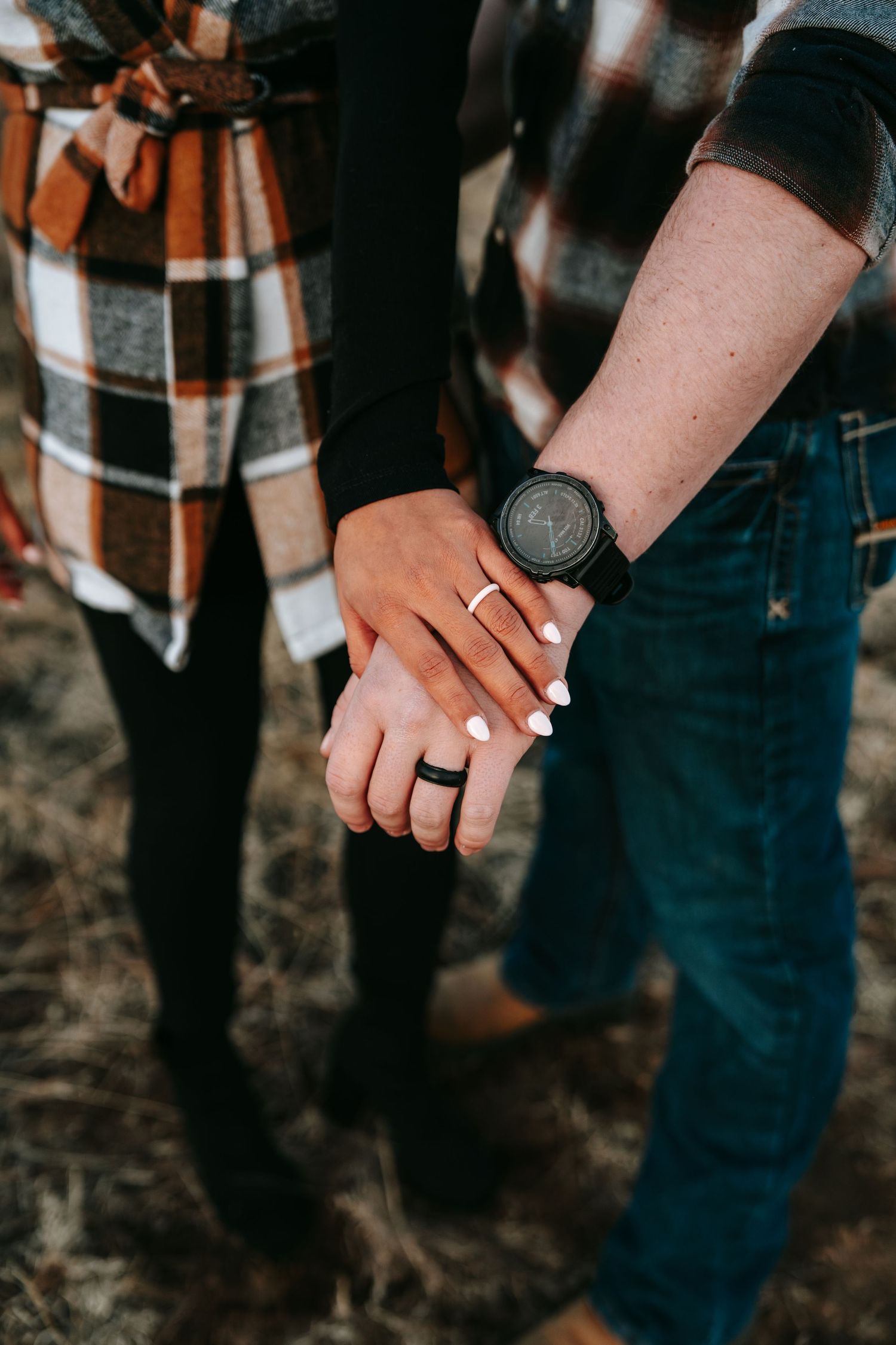 Couple holding hands outdoors, woman wearing ring and man wearing black watch, both in plaid flannel shirts.