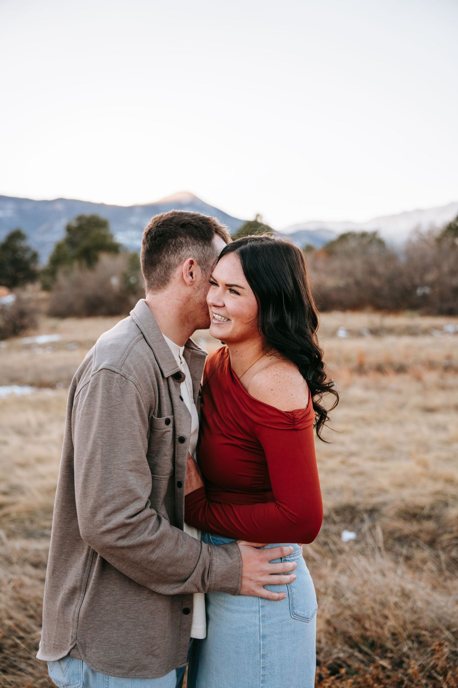 Man kissing smiling woman in red off-shoulder top in a golden winter landscape.