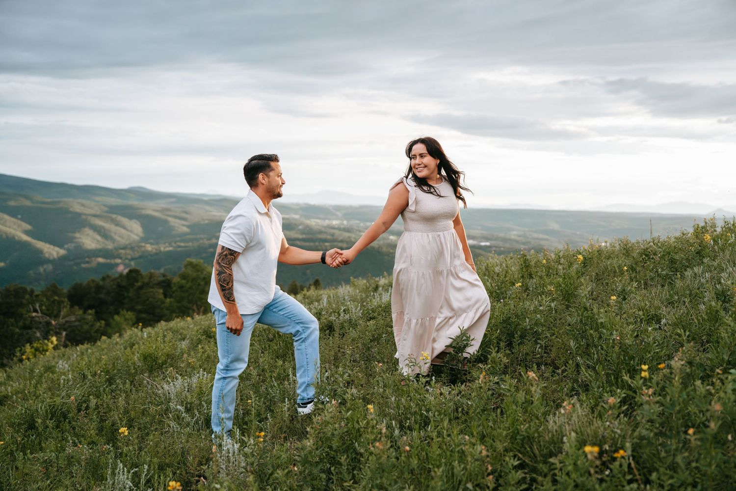 Couple holds hands and walks through wildflower meadow with scenic mountain valley behind them.