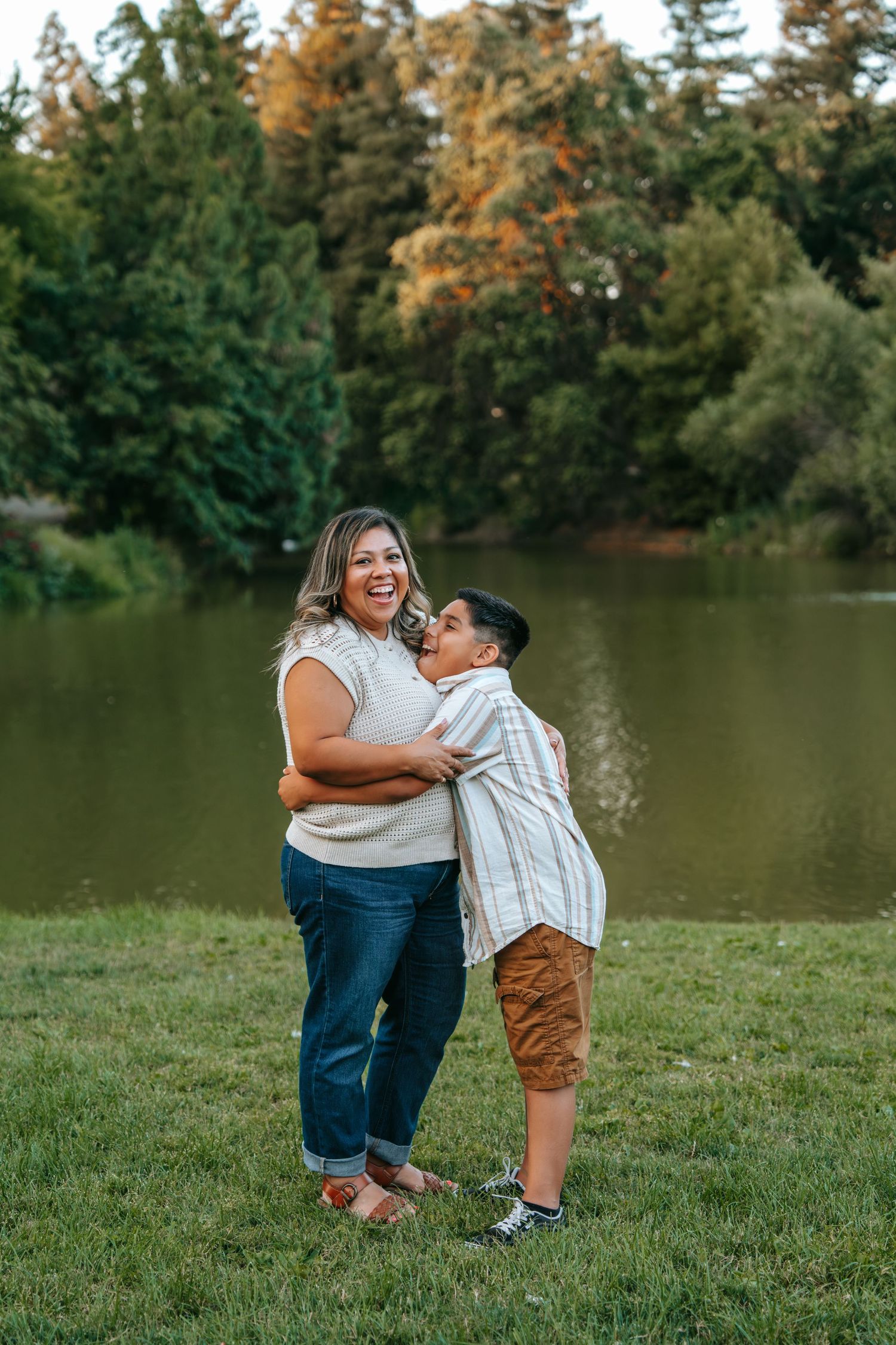 Smiling mother laughing as her son gives her a kiss on the cheek by a park pond.