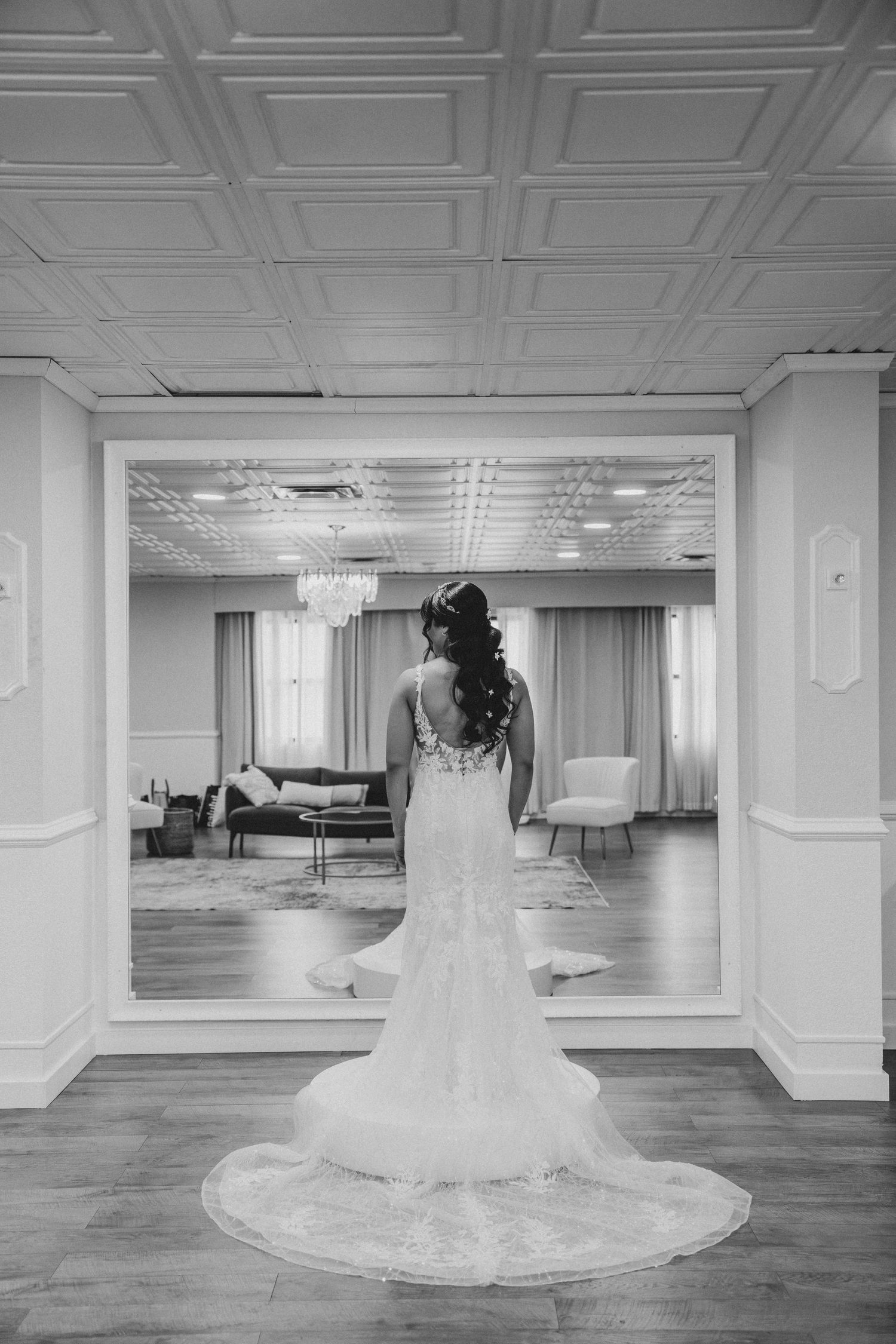 Bride in lace mermaid wedding gown with long train stands before mirror in elegant black and white bridal suite photo.