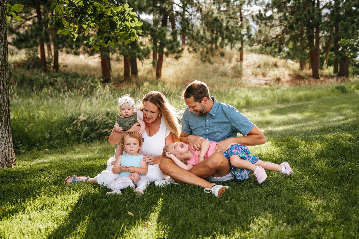 Happy family of five sitting on lush green grass in a sunny park, parents playing with three young children.