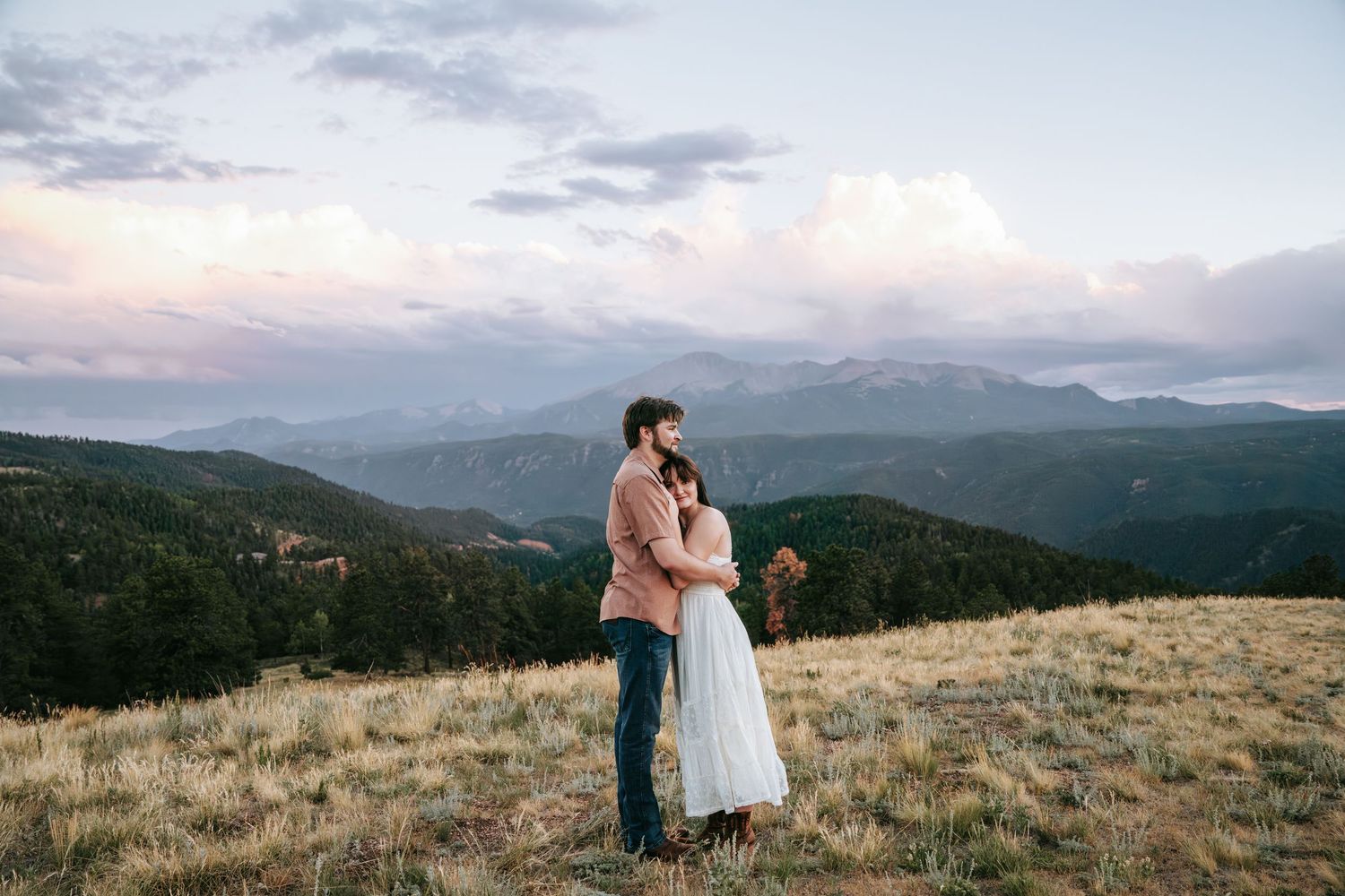 Couple embracing on a mountain meadow at sunset with scenic Rocky Mountain views in the background.