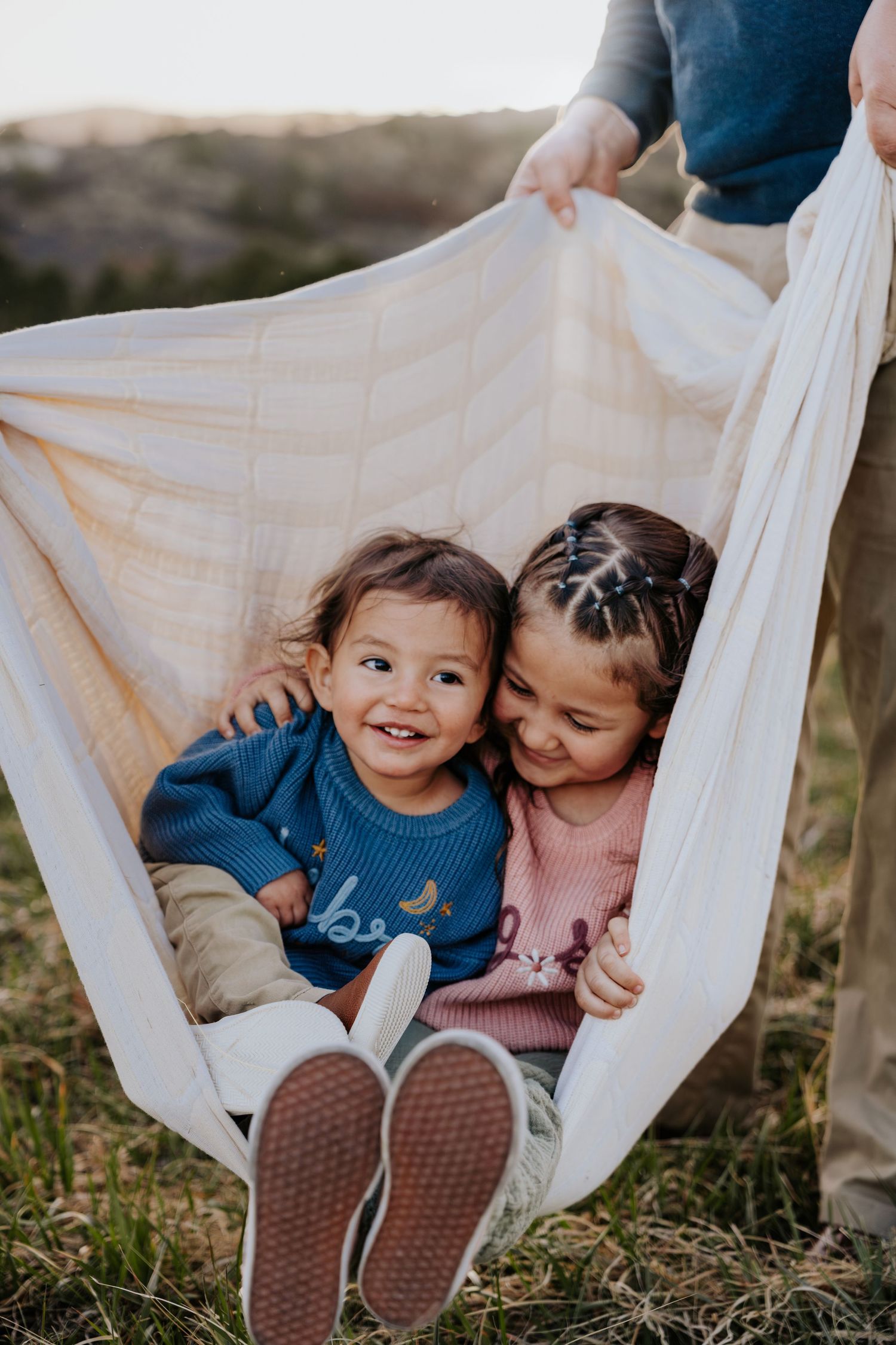 Two young siblings smile and cuddle together while being held in a swinging blanket outdoors at sunset.