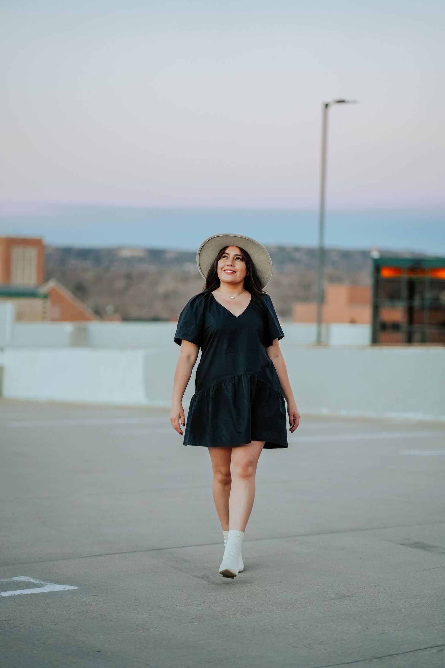 A young woman in a black mini dress and tan hat strolls across a rooftop at sunset.