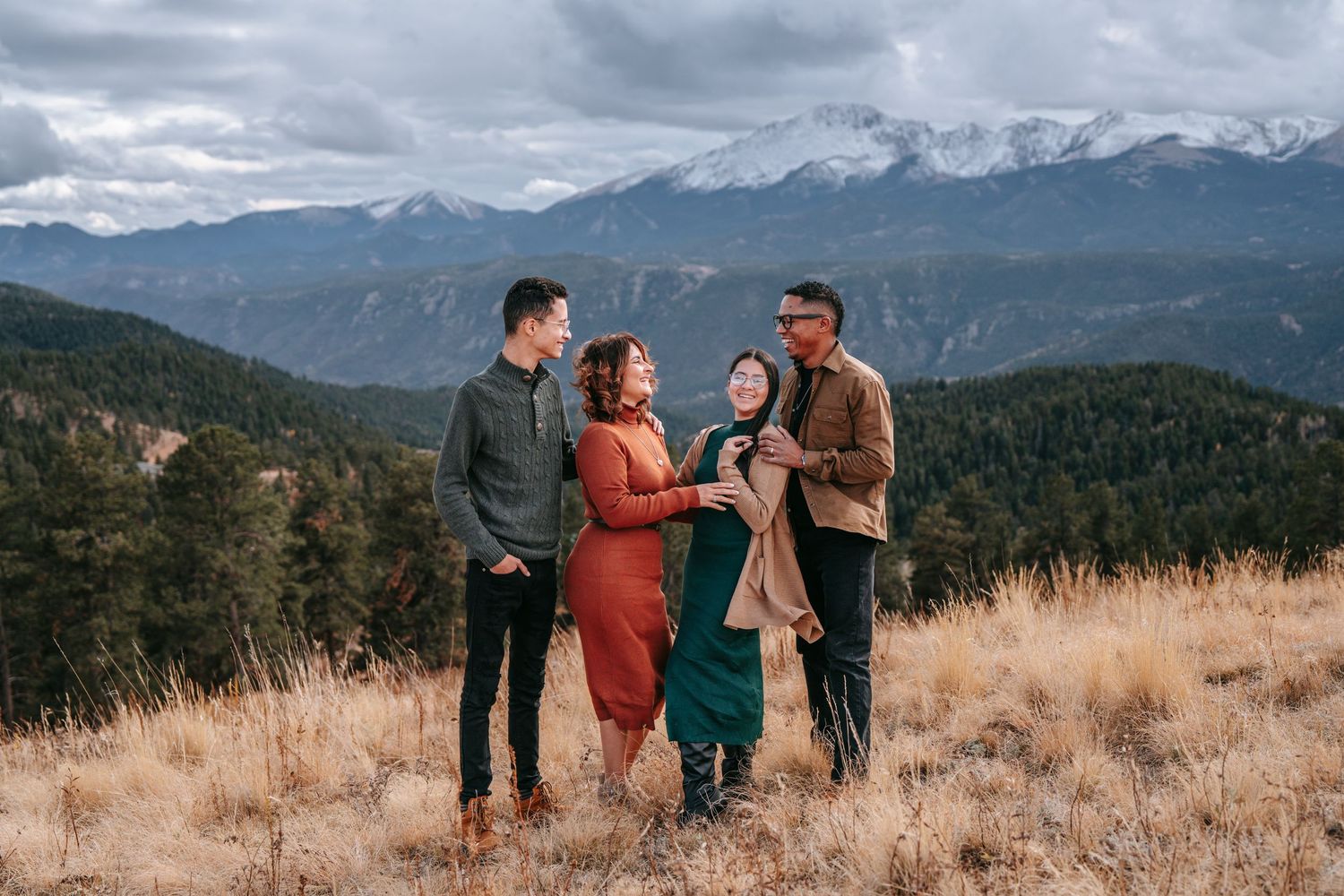 Four diverse friends laughing together in a mountain meadow with snow-capped peaks and pine trees behind them.