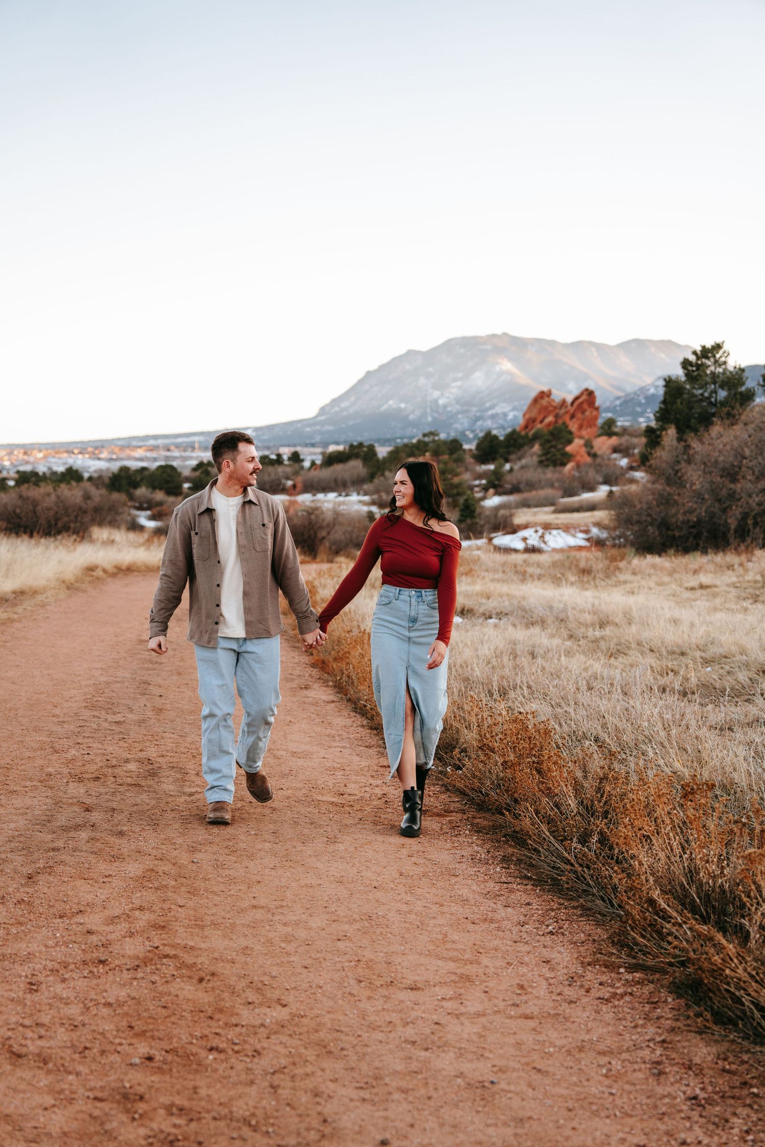 Couple holding hands walking on a red dirt trail with Garden of the Gods in background.