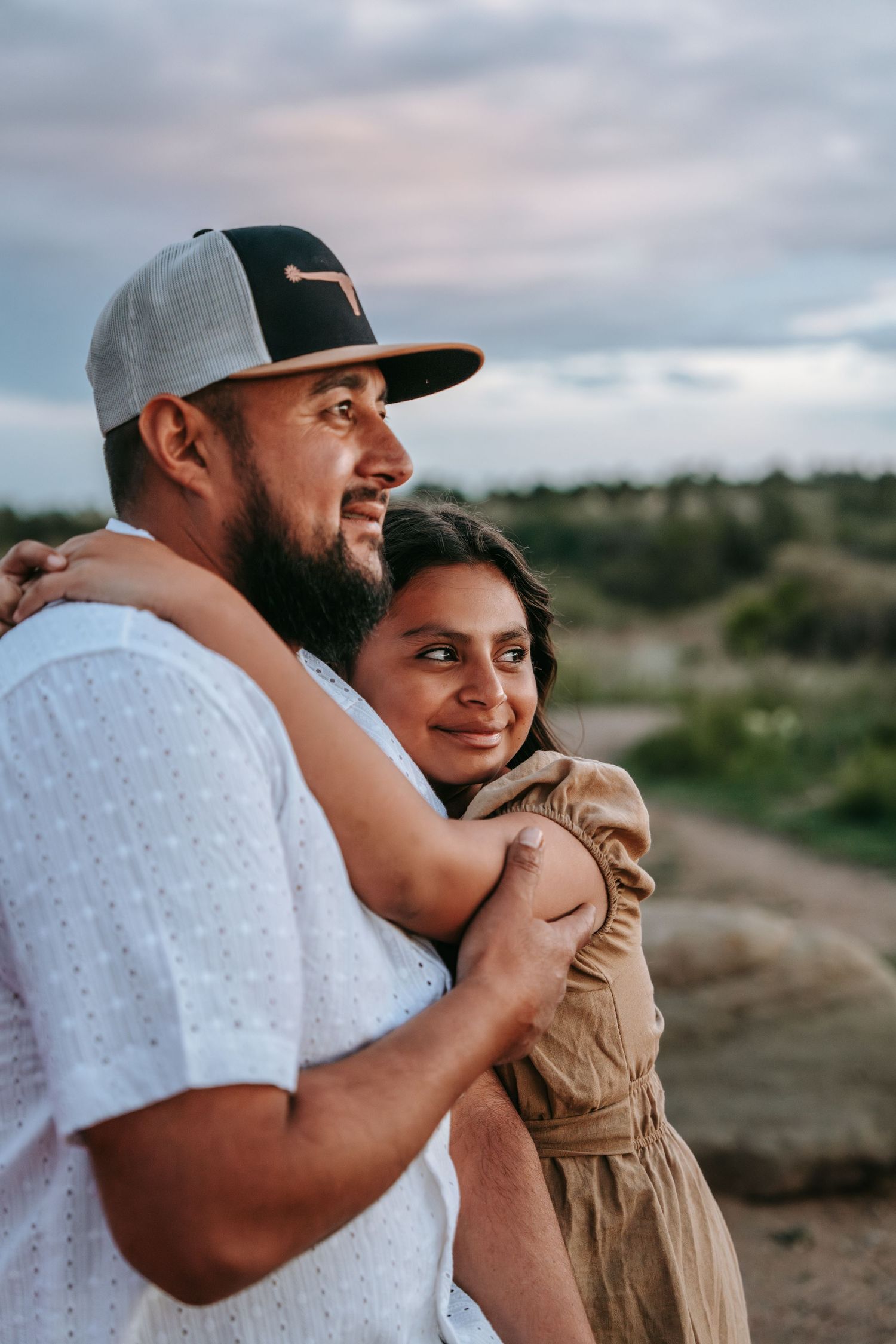 Bearded man in cap and woman in tan dress embrace while gazing at scenic outdoor landscape.
